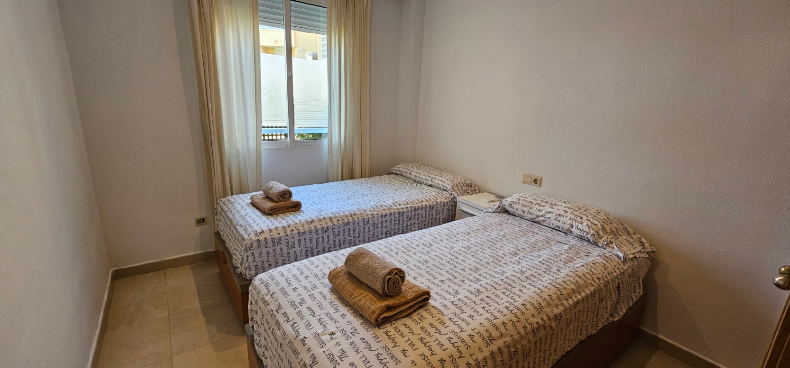 Slaapkamer voorzien van twee eenpersoonsbedden, een lichte tegelvloer en een raam met witte gordijnen.