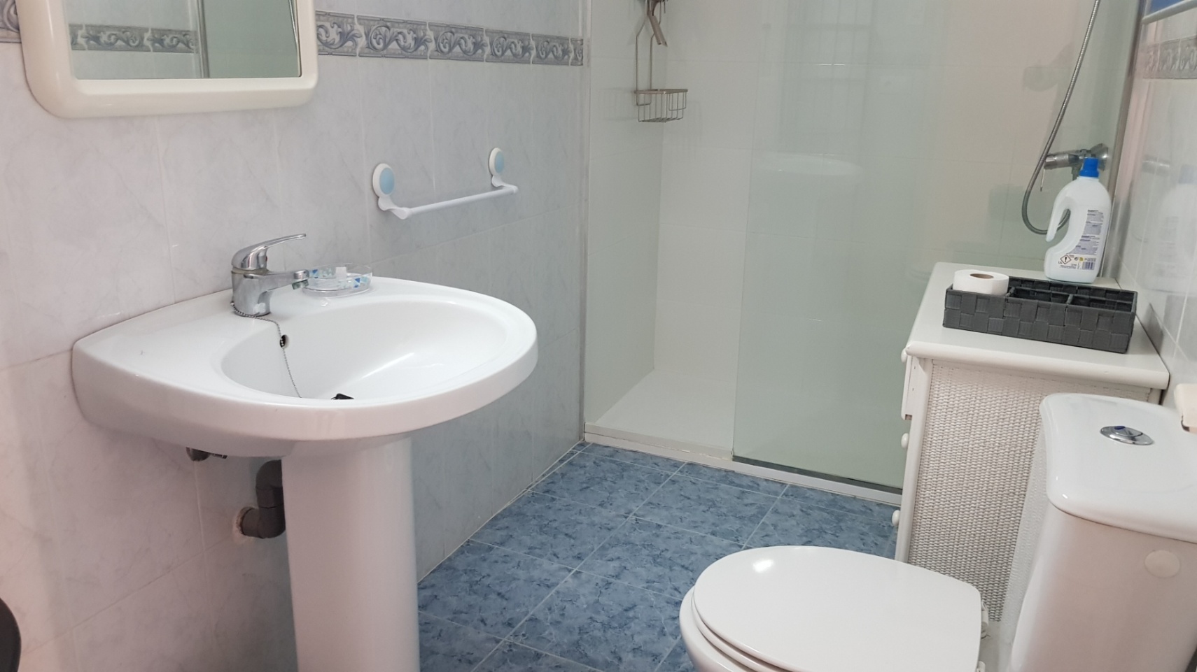 Badkamer met blauwe vloertegels, een wastafel op zuil en een douche met glazen wand. Witte wandtegels met een decoratieve rand sieren de ruimte.