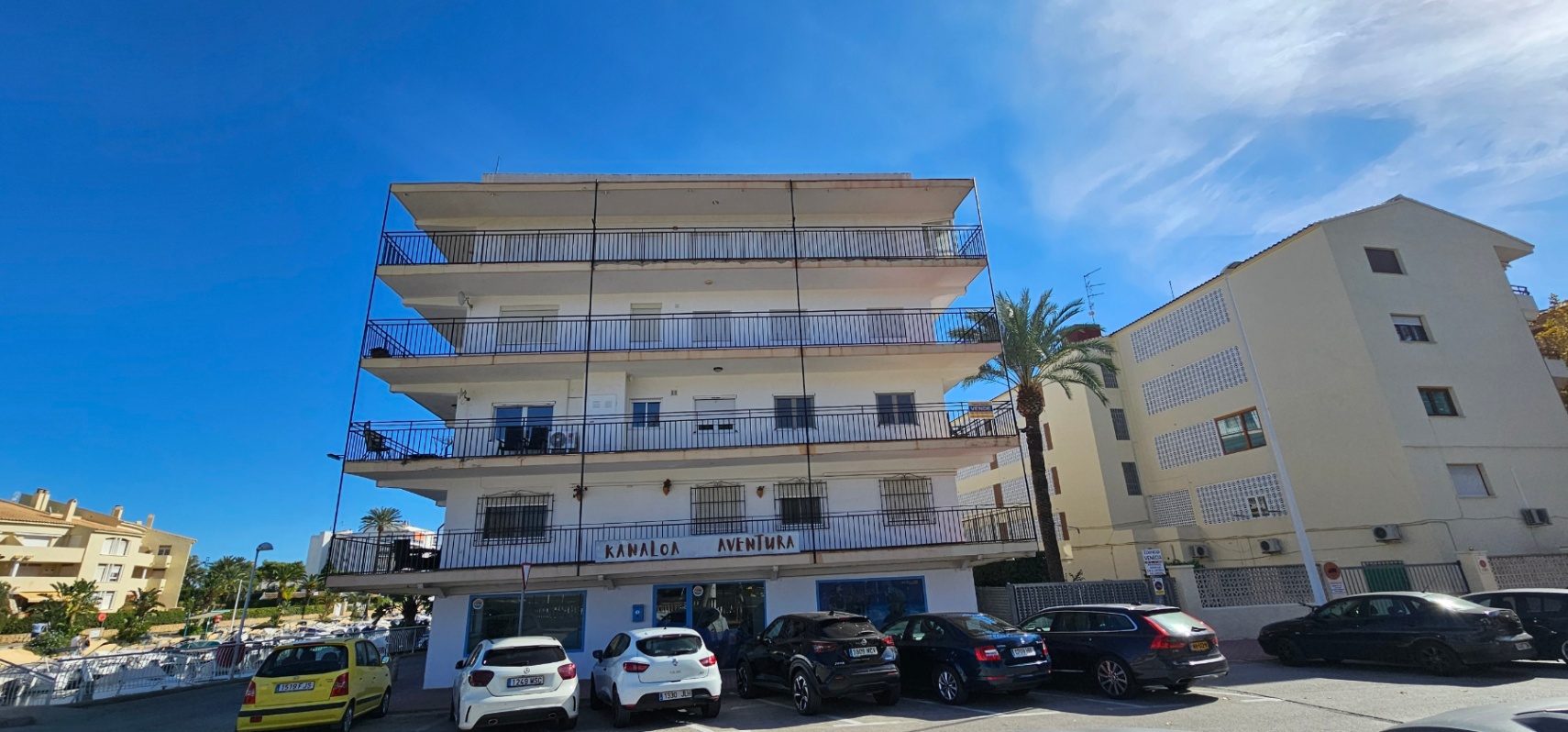 Buitenaanzicht van het gebouw in mediterrane stijl met doorlopende balkons, zwart metalen hekwerk en een commerciële ruimte op de begane grond.