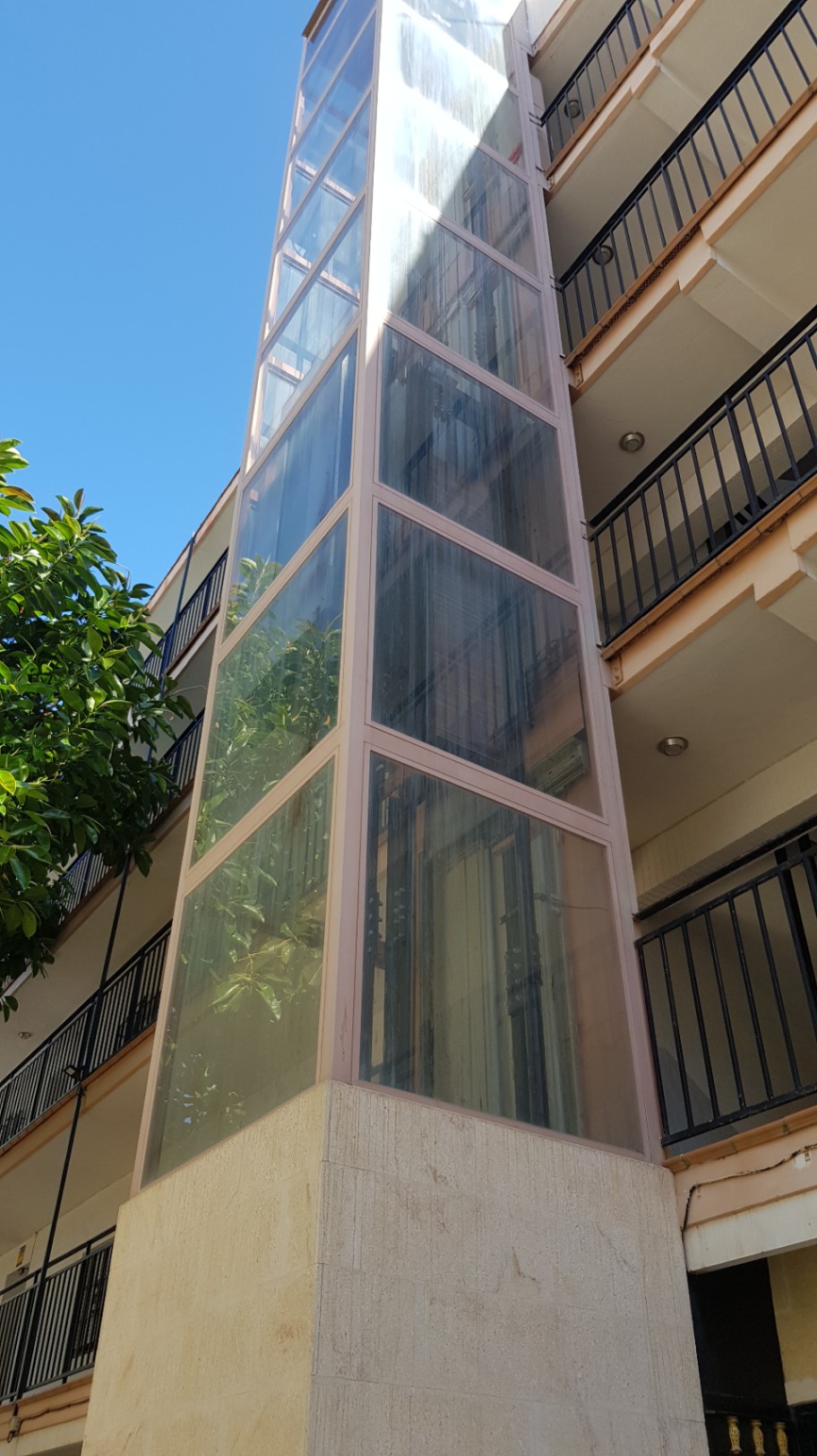 Externe panoramische glazen lift met een stenen basis, geïntegreerd in de gevel van het woongebouw naast balkons met zwarte metalen leuningen.