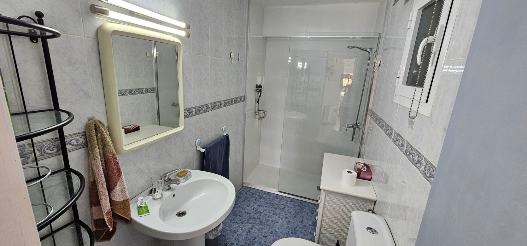 Functionele badkamer met inloopdouche, glazen wand, kolomwastafel en blauwe vloertegels gecombineerd met een klassieke wandafwerking.