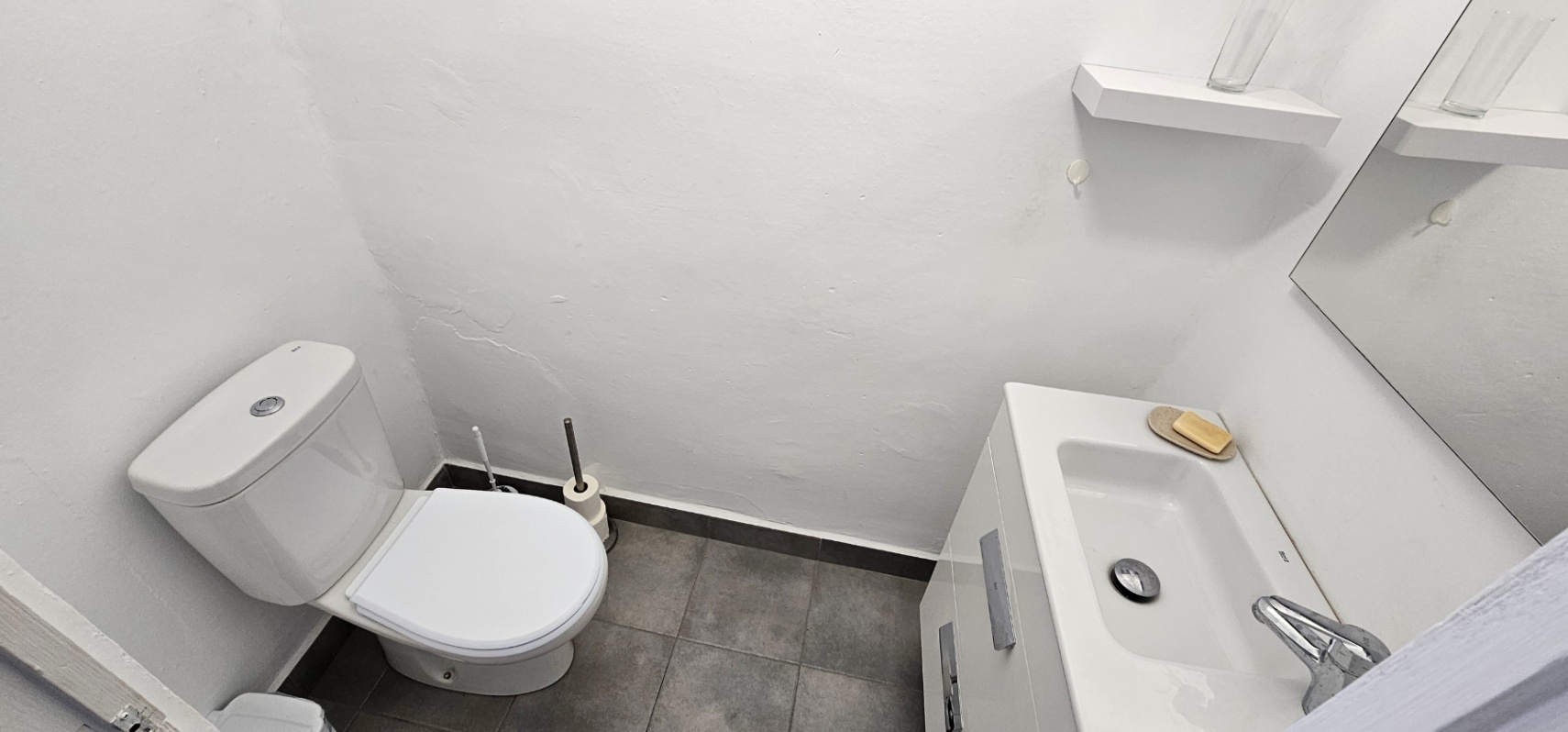 Functionele badkamer met wit toilet, wastafelmeubel met geïntegreerde wasbak en grijze vloertegels. Inclusief een spiegel en een minimalistische wandplank.