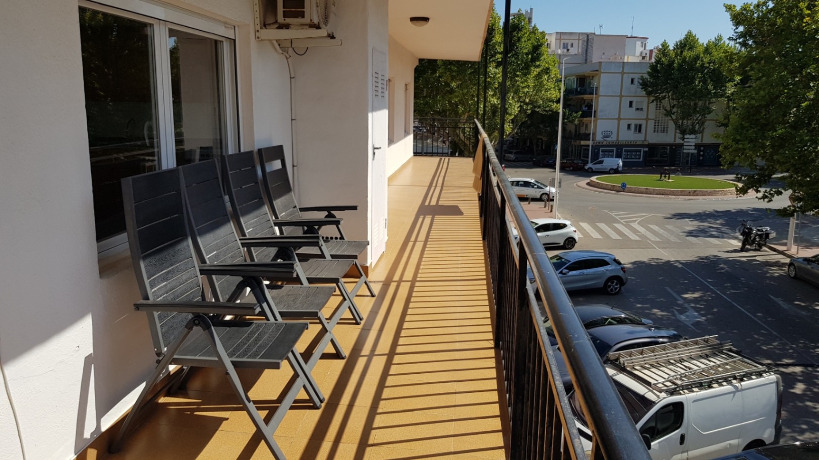 Lang balkon met okergele vloertegels, zwart metalen balustrade en inklapbare buitenstoelen met uitzicht op de straat en de groene omgeving van de stad.