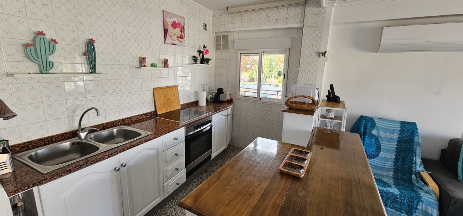 Open keuken met granieten werkblad, dubbele spoelbak en witte kasten. Voorzien van een houten eettafel, airconditioning en een raam met natuurlijk licht.