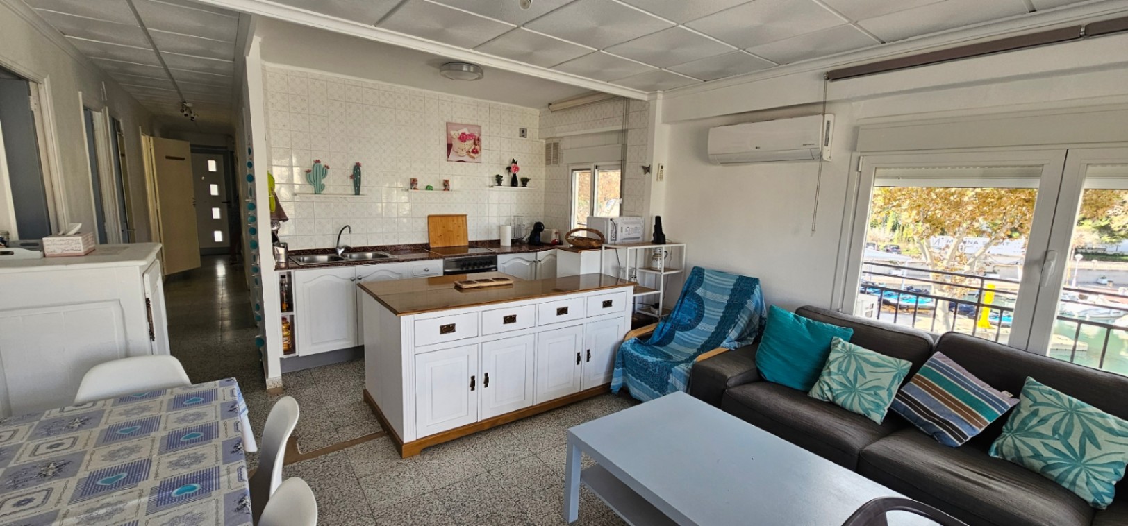 Open woonkamer met geïntegreerde keuken, houten kookeiland en directe toegang tot het terras met uitzicht op de jachthaven en boten.