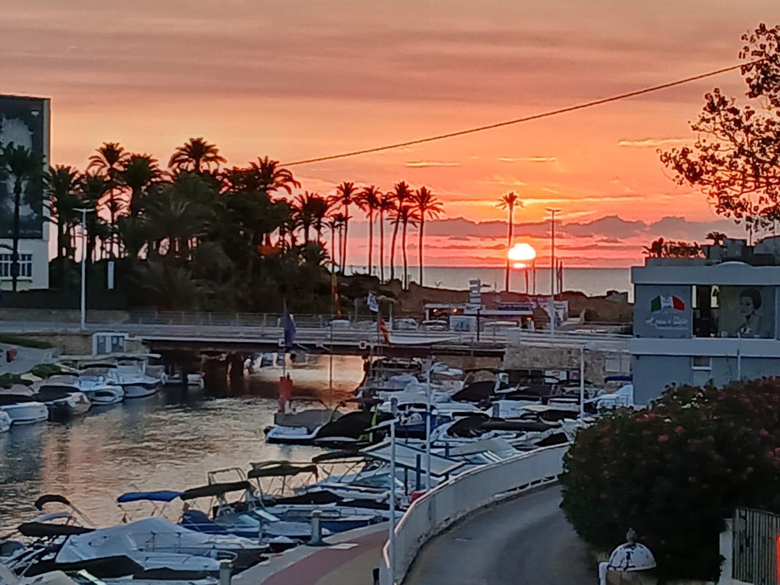 Panoramisch uitzicht op de jachthaven en het kanaal bij zonsondergang, met aangemeerde boten en silhouetten van palmbomen tegen een oranje lucht aan de horizon.