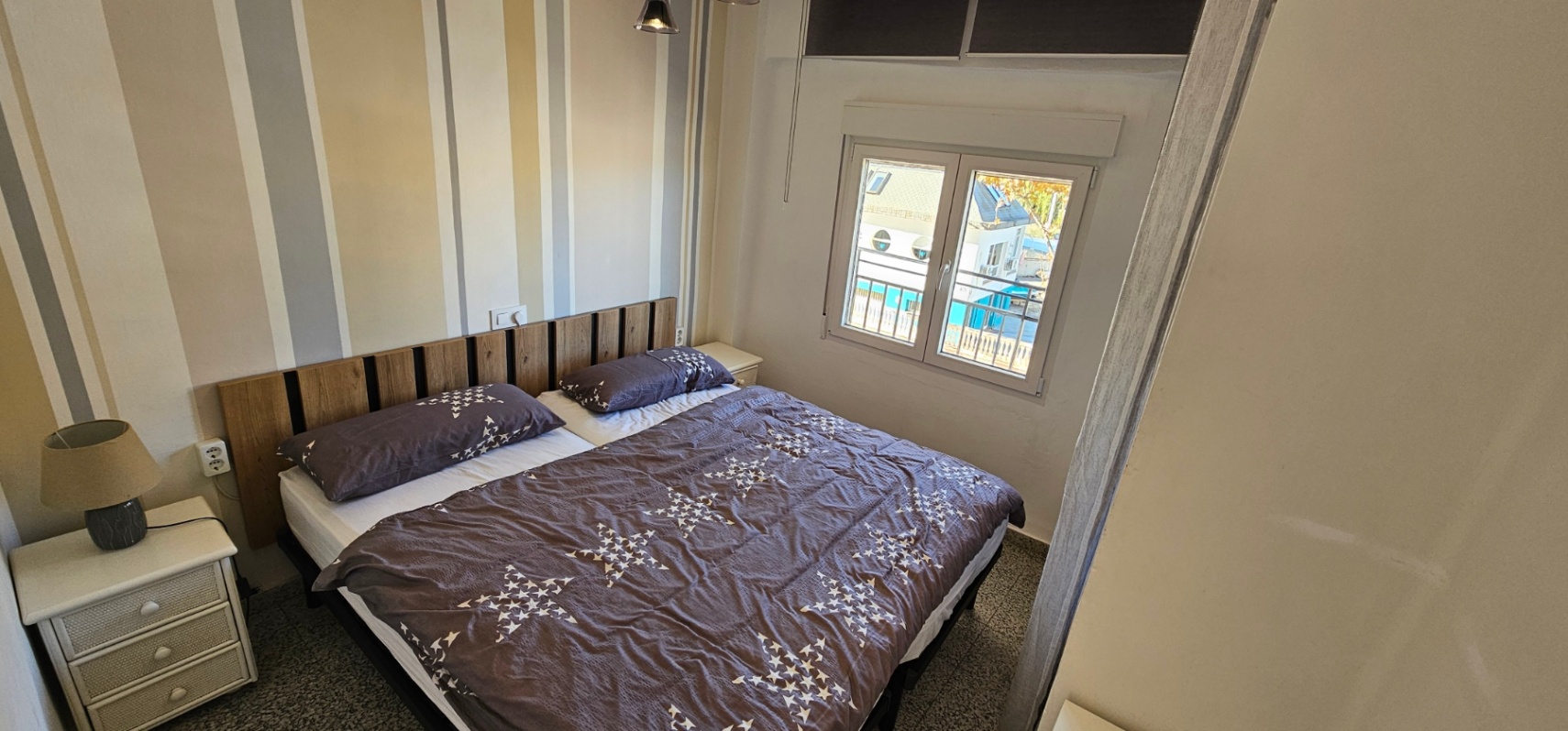 Slaapkamer met tweepersoonsbed, houten latten hoofdbord en gestreept behang. Het raam zorgt voor natuurlijk licht en biedt uitzicht op de nabijgelegen jachthaven.
