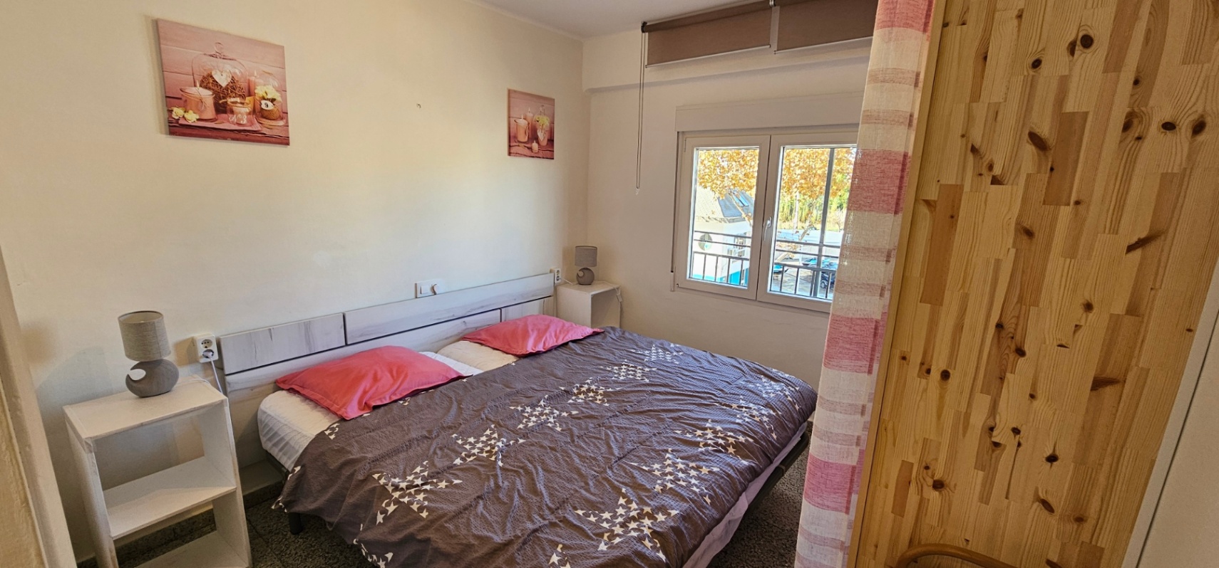 Slaapkamer met tweepersoonsbed, licht houten hoofdbord en witte nachtkastjes. Een raam biedt natuurlijk licht en uitzicht op de bomen en de straat buiten.