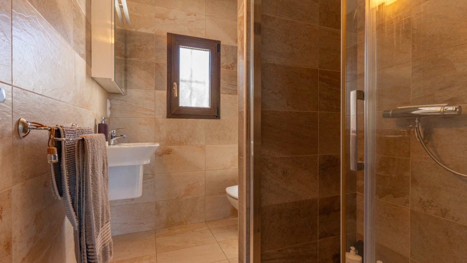 Badkamer met keramische tegels in steenlook, een glazen douchecabine, witte wastafel en een klein raam met houten kozijn.