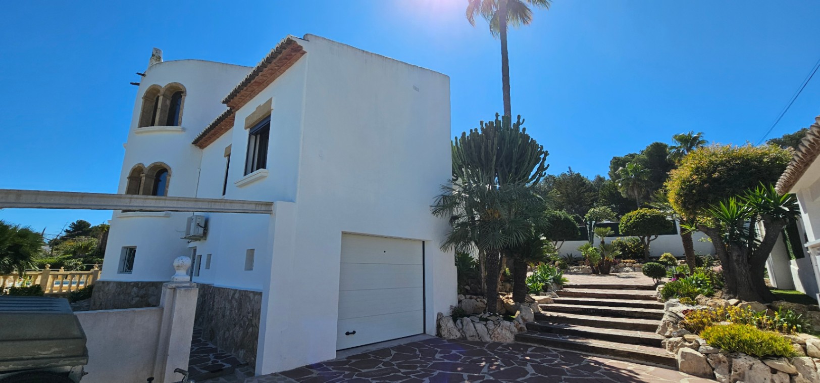 Buitenaanzicht van de witte villa met natuurstenen plint, geïntegreerde garage en stenen trap omgeven door mediterrane beplanting en palmbomen.