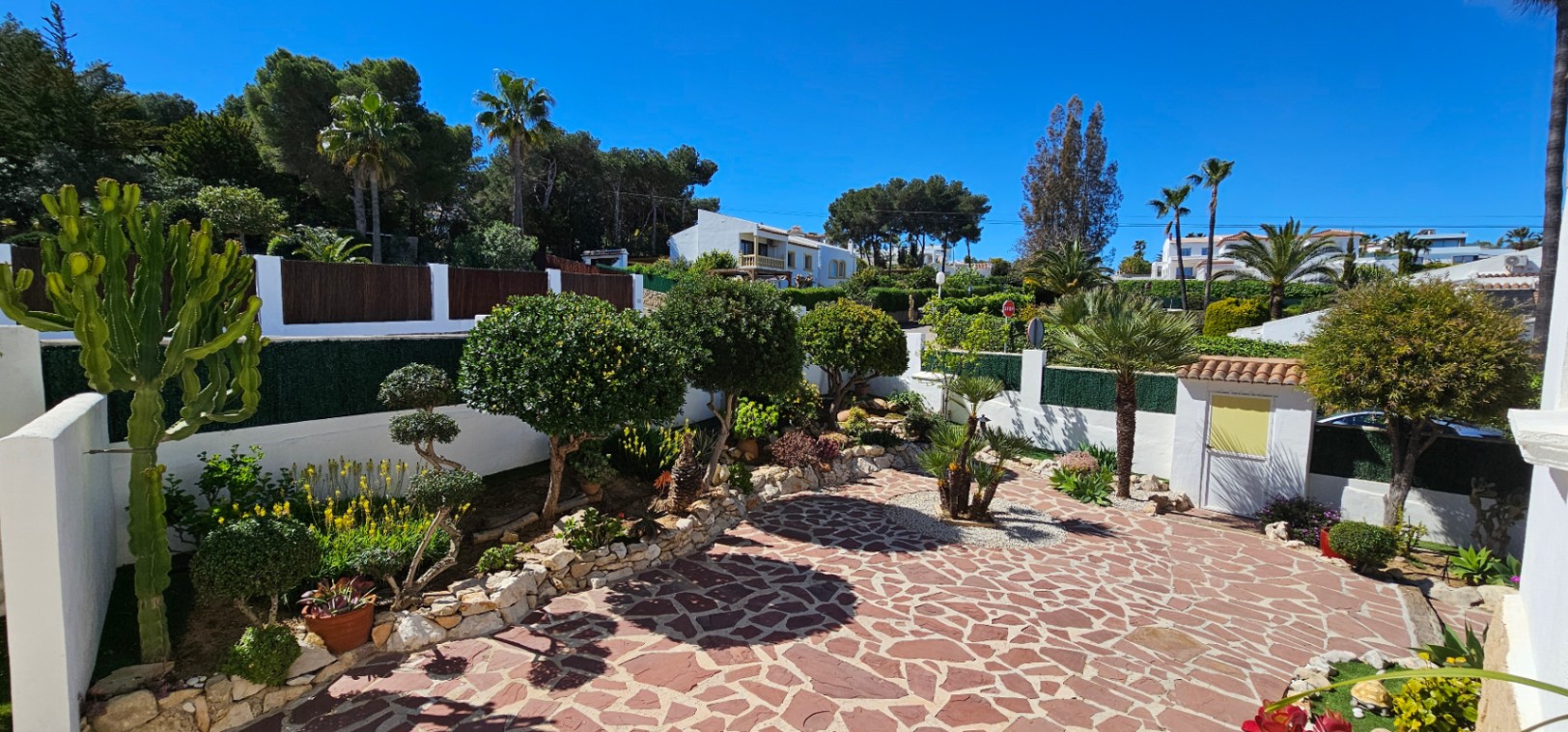 Buitenplaats met roodachtige natuurstenen bestrating, witte muren en een mediterrane tuin met palmen, cactussen en zorgvuldig gesnoeide struiken.