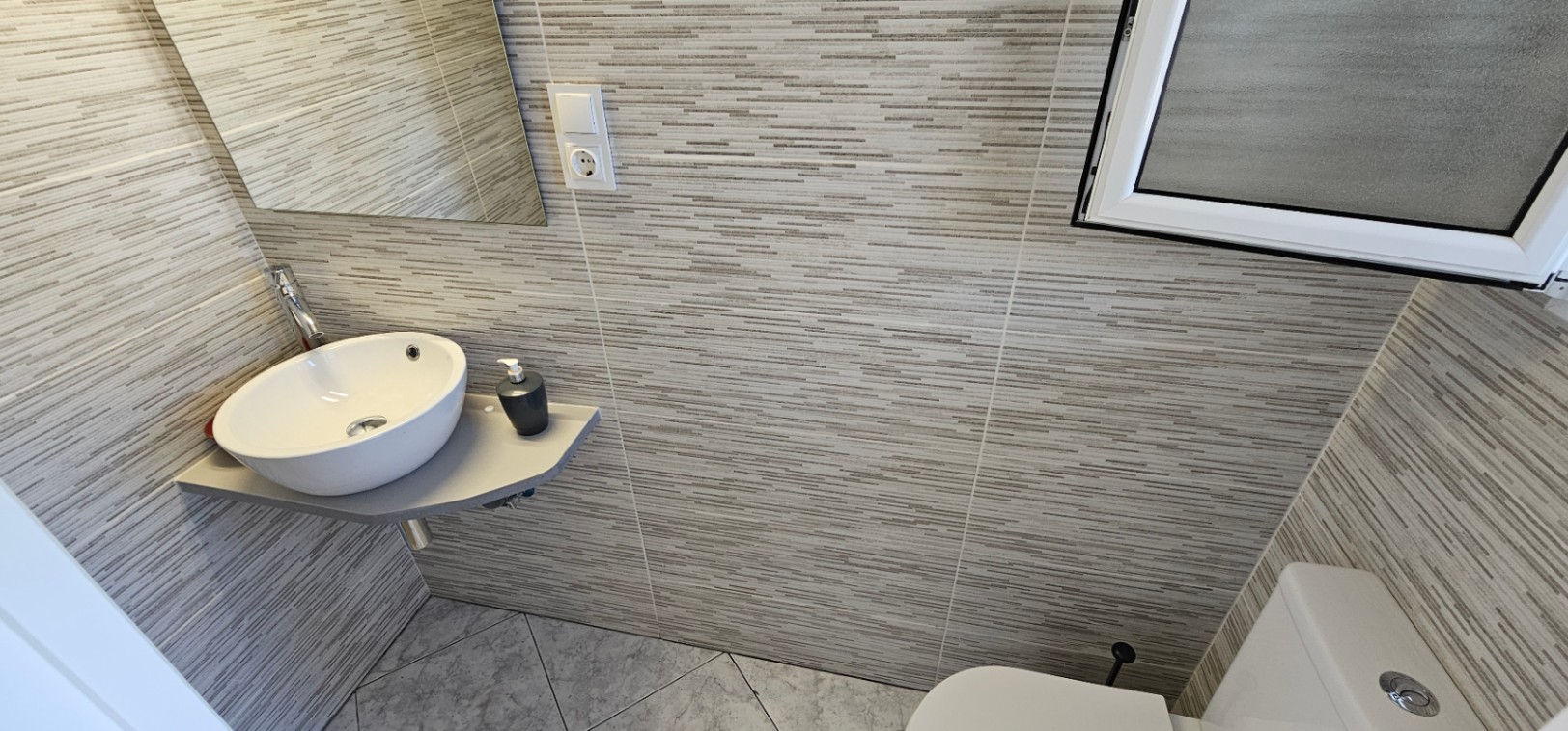 Compact gastentoilet met een hoekwastafel, horizontaal gestreepte grijze wandtegels en diagonaal gelegde keramische vloertegels.