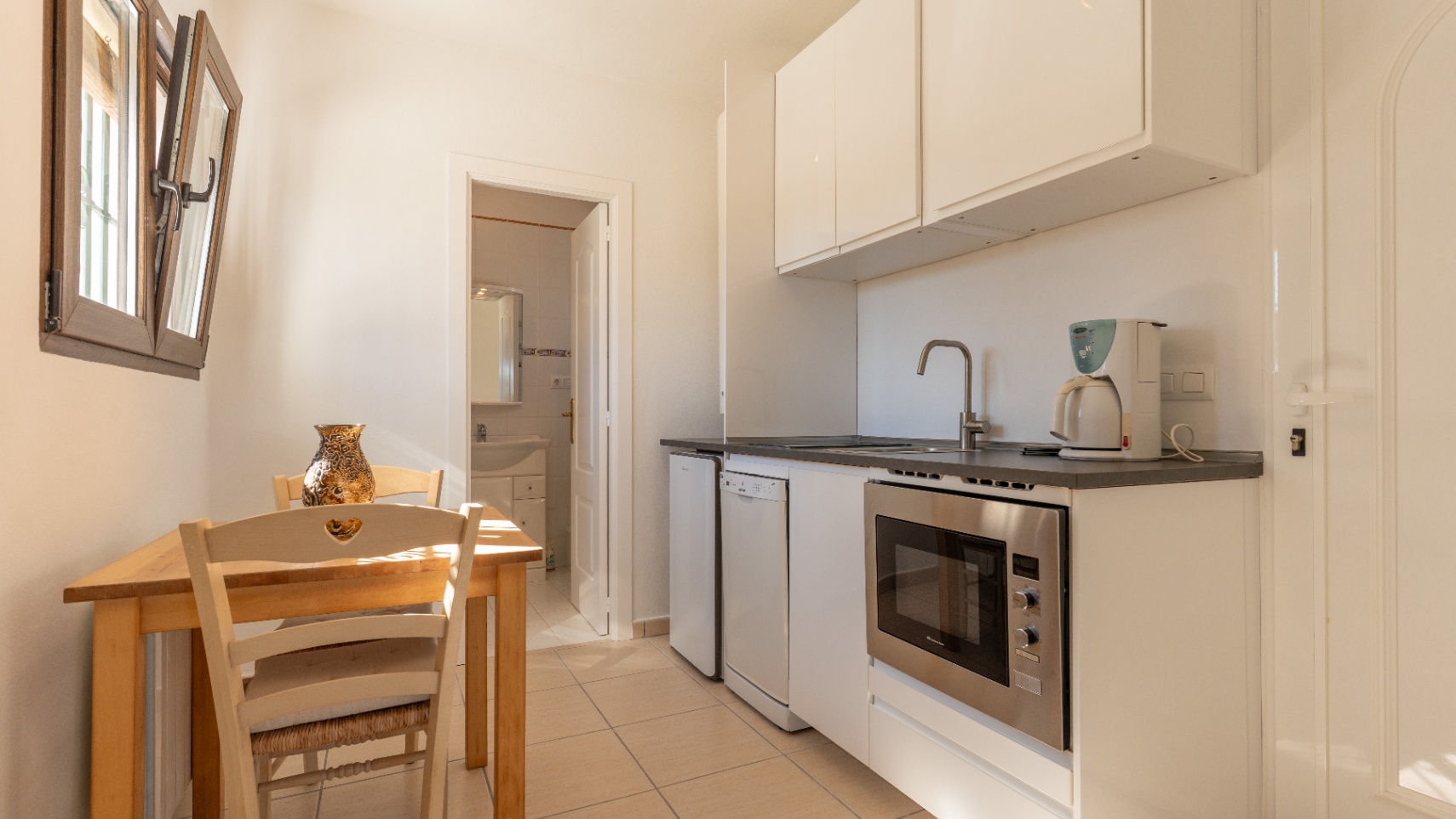 Compacte keuken met witte kasten, inbouwapparatuur en een houten eethoek, gelegen naast een aangrenzende badkamer.