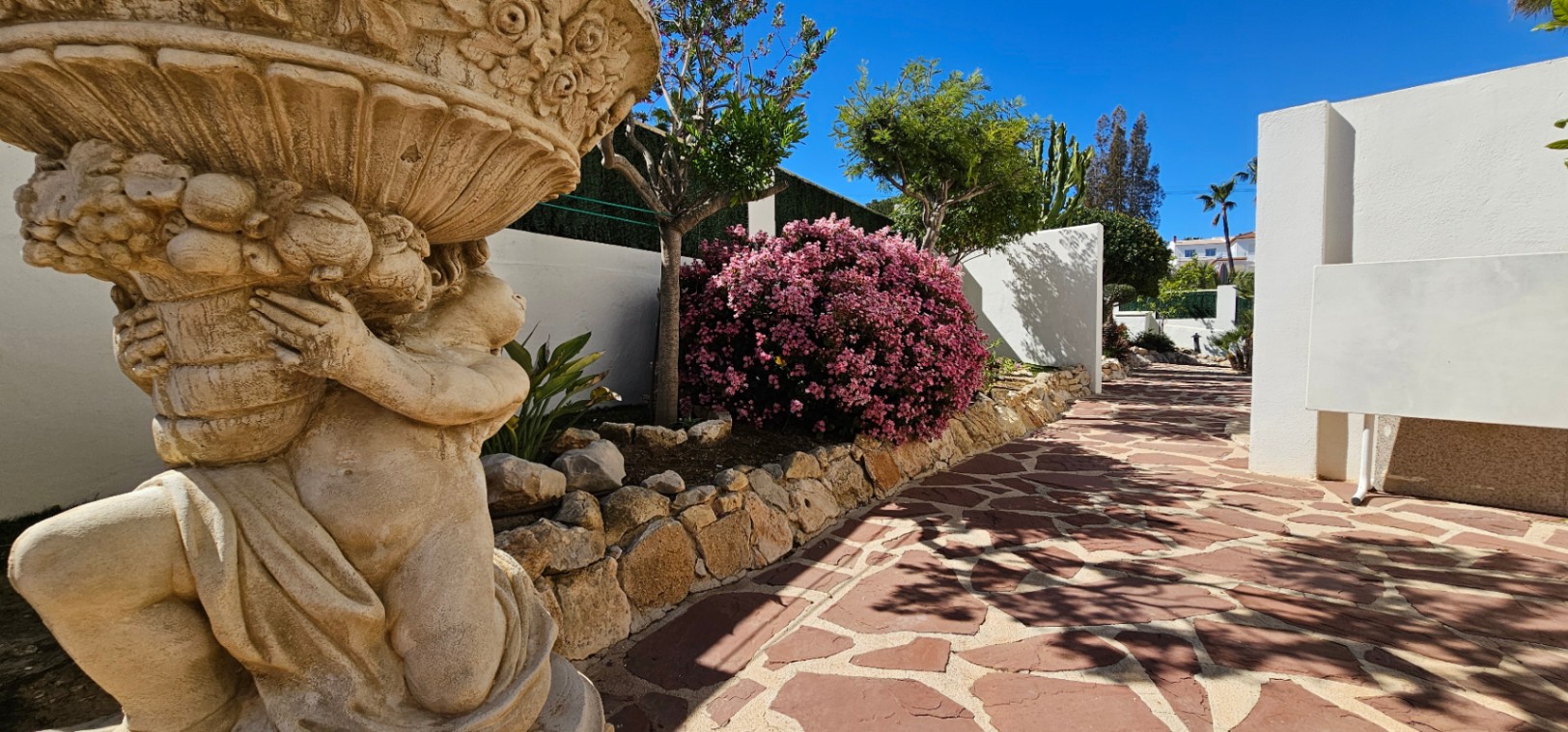 Een pad van natuursteen omgeven door een decoratief standbeeld en mediterrane beplanting, waaronder bloeiende roze struiken en witte muren.