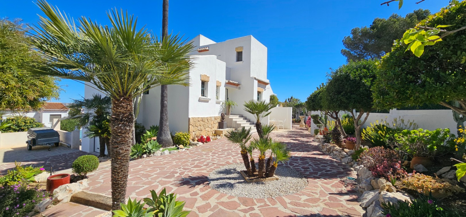 Exterieur van de villa met witte gevel en natuurstenen plint, omgeven door een mediterrane tuin met palmbomen en onregelmatige stenen bestrating.