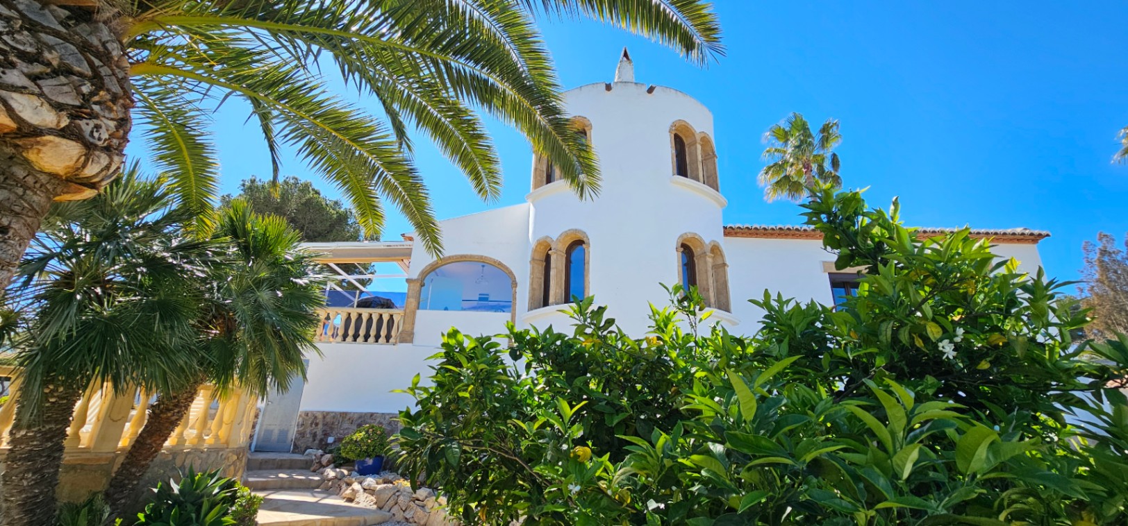 Exterieur van een mediterrane villa met een witte ronde toren, Tosca-stenen bogen en een weelderige tuin met palmbomen en citrusvruchten onder een strakblauwe lucht.