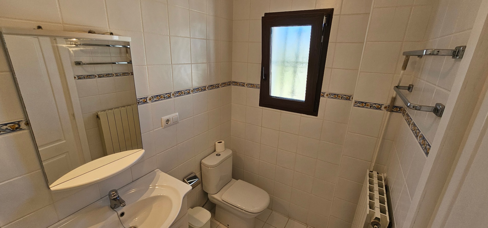 Functionele badkamer met witte wandtegels en een decoratieve rand. Voorzien van een keramische wastafel, radiator en een raam met melkglas voor privacy.
