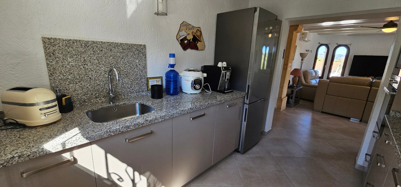 Functionele keuken met granieten aanrechtblad en rvs spoelbak, grenzend aan een lichte woonkamer met lederen banken en karakteristieke boogramen.