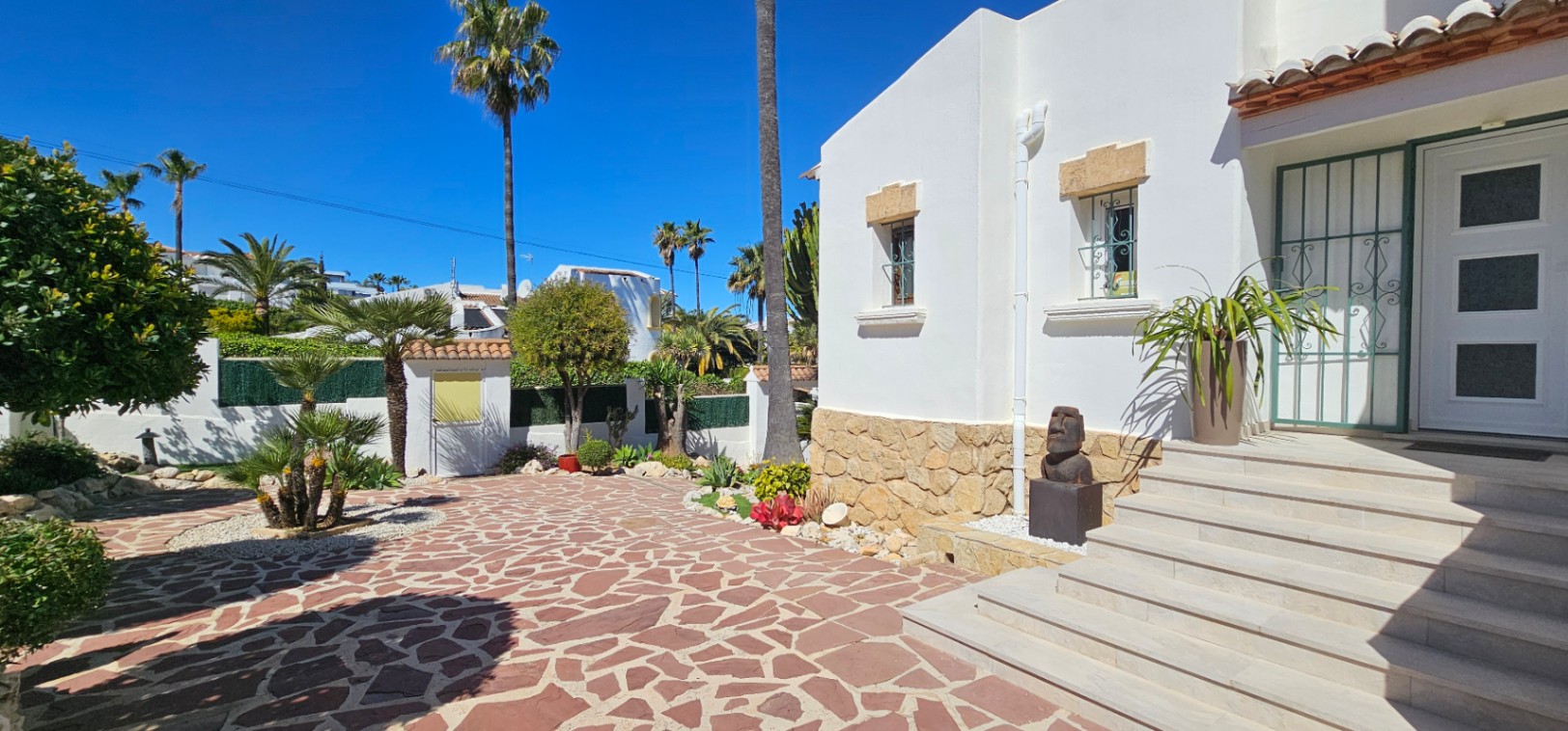Hoofdingang met brede witte treden en een mediterrane gevel. De buitenruimte is voorzien van onregelmatige stenen bestrating en een aangelegde tuin met palmbomen.