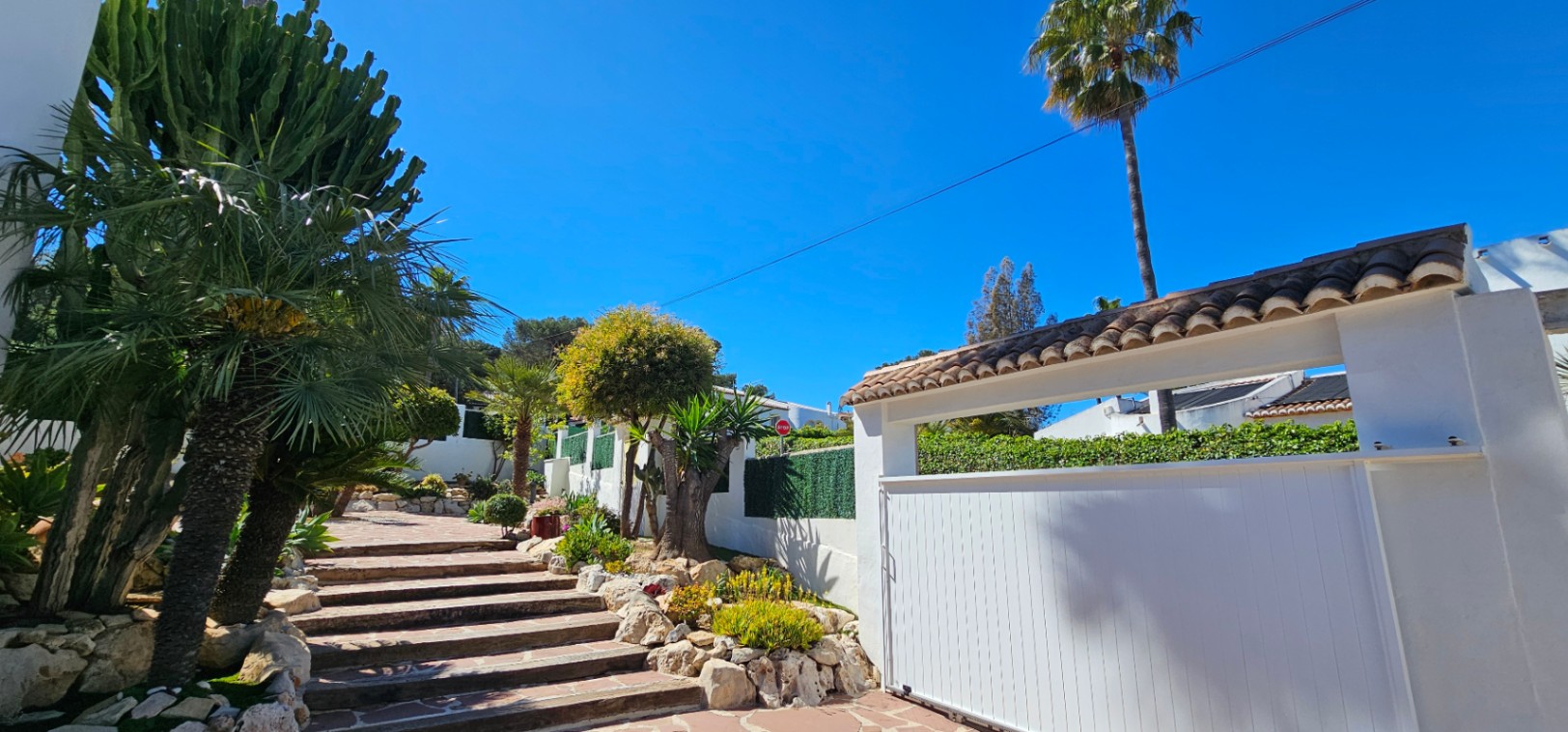 Hoofdingang met een witte schuifpoort en stenen trap, omgeven door een mediterrane tuin met cactussen en palmbomen tegen een helderblauwe lucht.