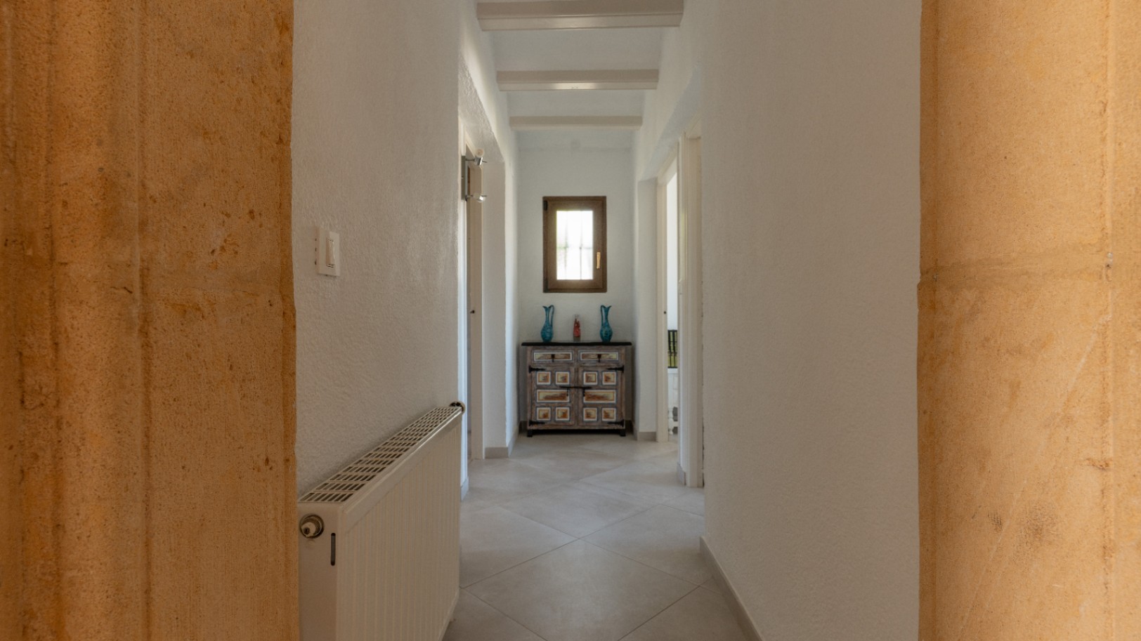 Lichte gang met witte plafondbalken en Tosca natuurstenen bogen. Voorzien van centrale verwarming en een decoratief dressoir onder een houten raam.
