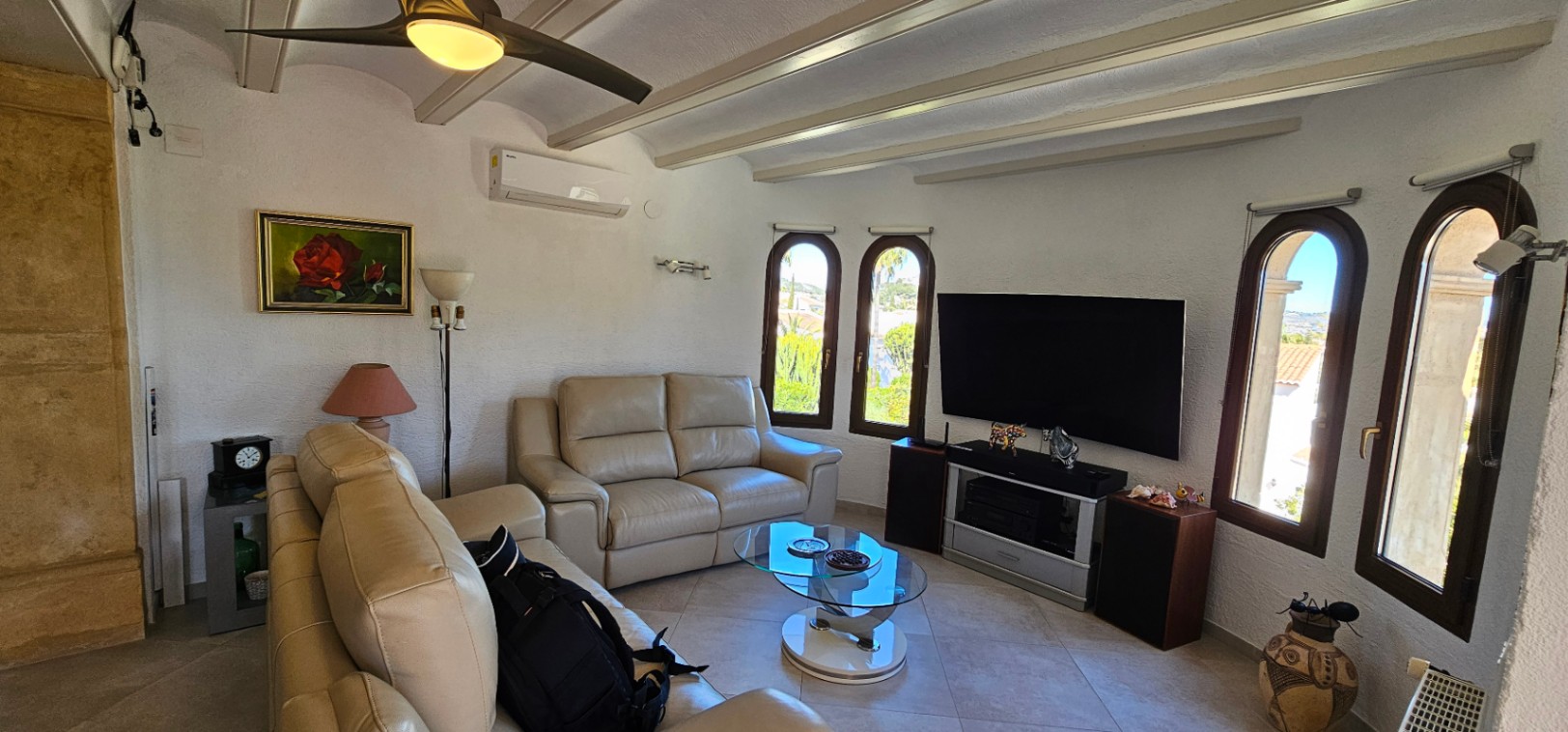 Lichte woonkamer met beige lederen banken, zichtbare plafondbalken, airconditioning en boogvensters die zorgen voor een overvloed aan natuurlijk licht.