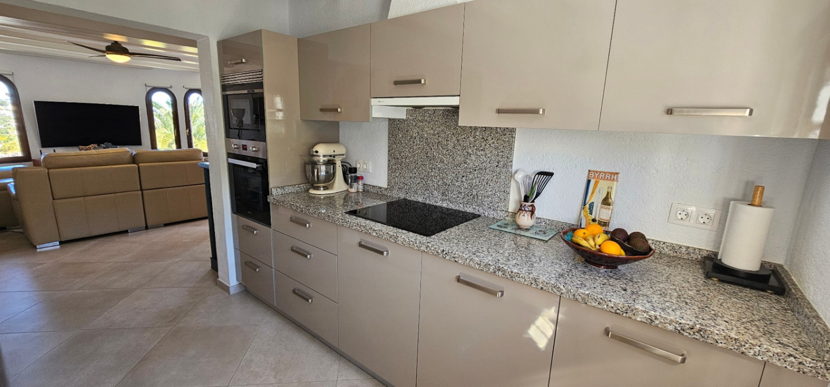 Moderne keuken met granieten werkbladen, hoogglans kasten in taupe en inbouwapparatuur, grenzend aan een lichte woonkamer met karakteristieke boogvensters.