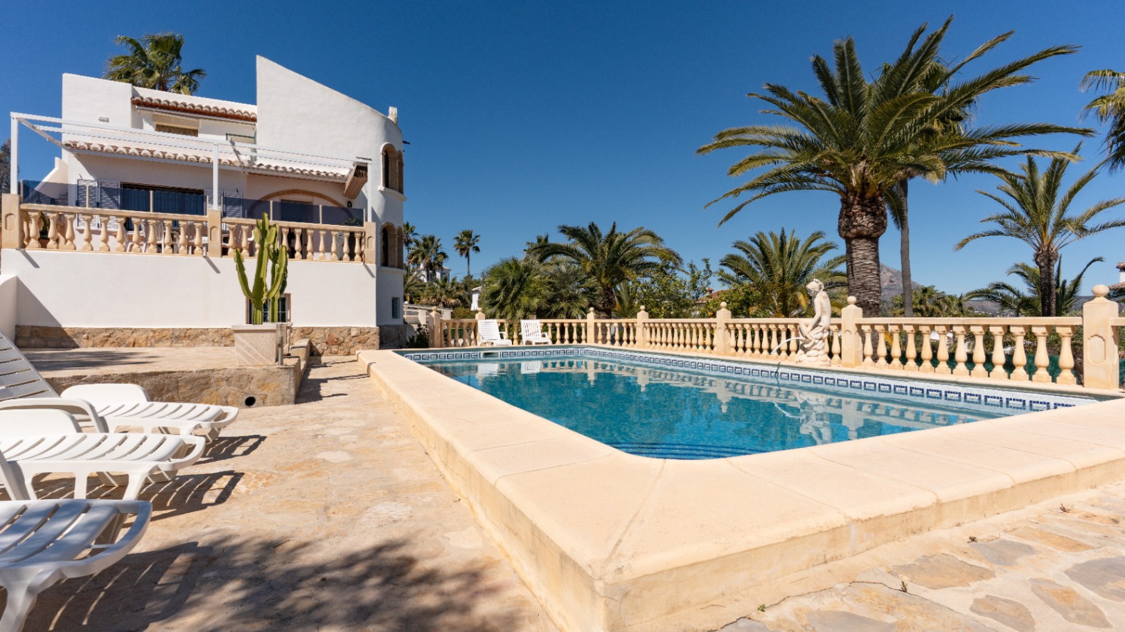 Rechthoekig zwembad met een ruim stenen zonneterras en ligstoelen, omgeven door volwassen palmbomen en de witte mediterrane gevel van de villa.