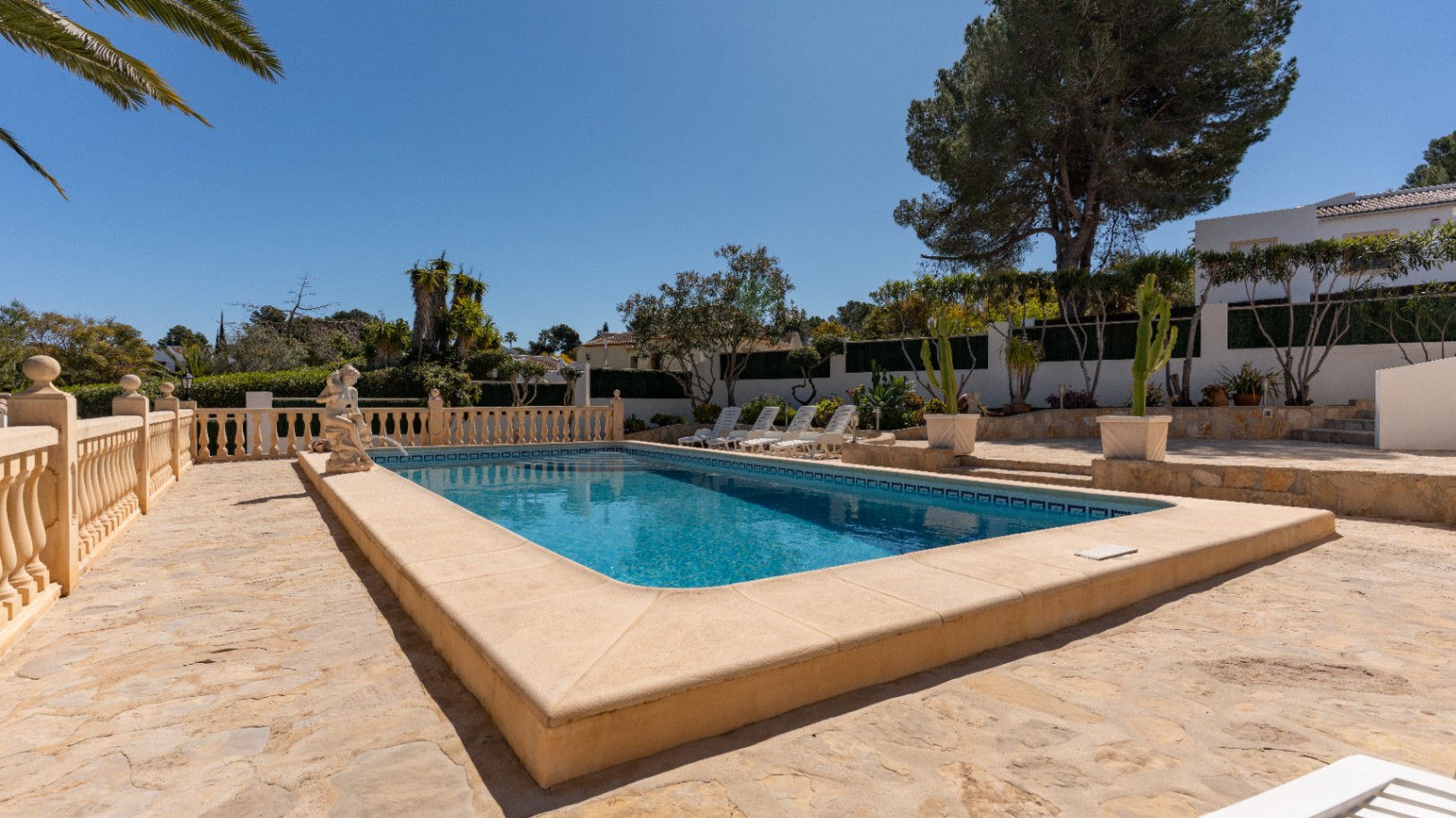 Rechthoekig zwembad omgeven door een natuurstenen terras met klassieke balustrade, decoratief beeld en zonneweide te midden van mediterrane beplanting.