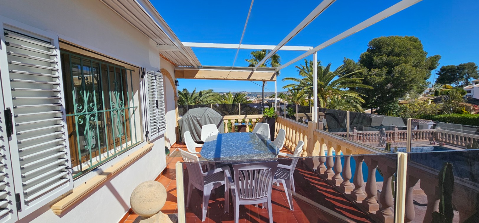 Verhoogd terras met terracotta vloer, granieten tafel en glazen balustrades. Biedt uitzicht op het zwembad en de omliggende palmbomen in een zonnige setting.