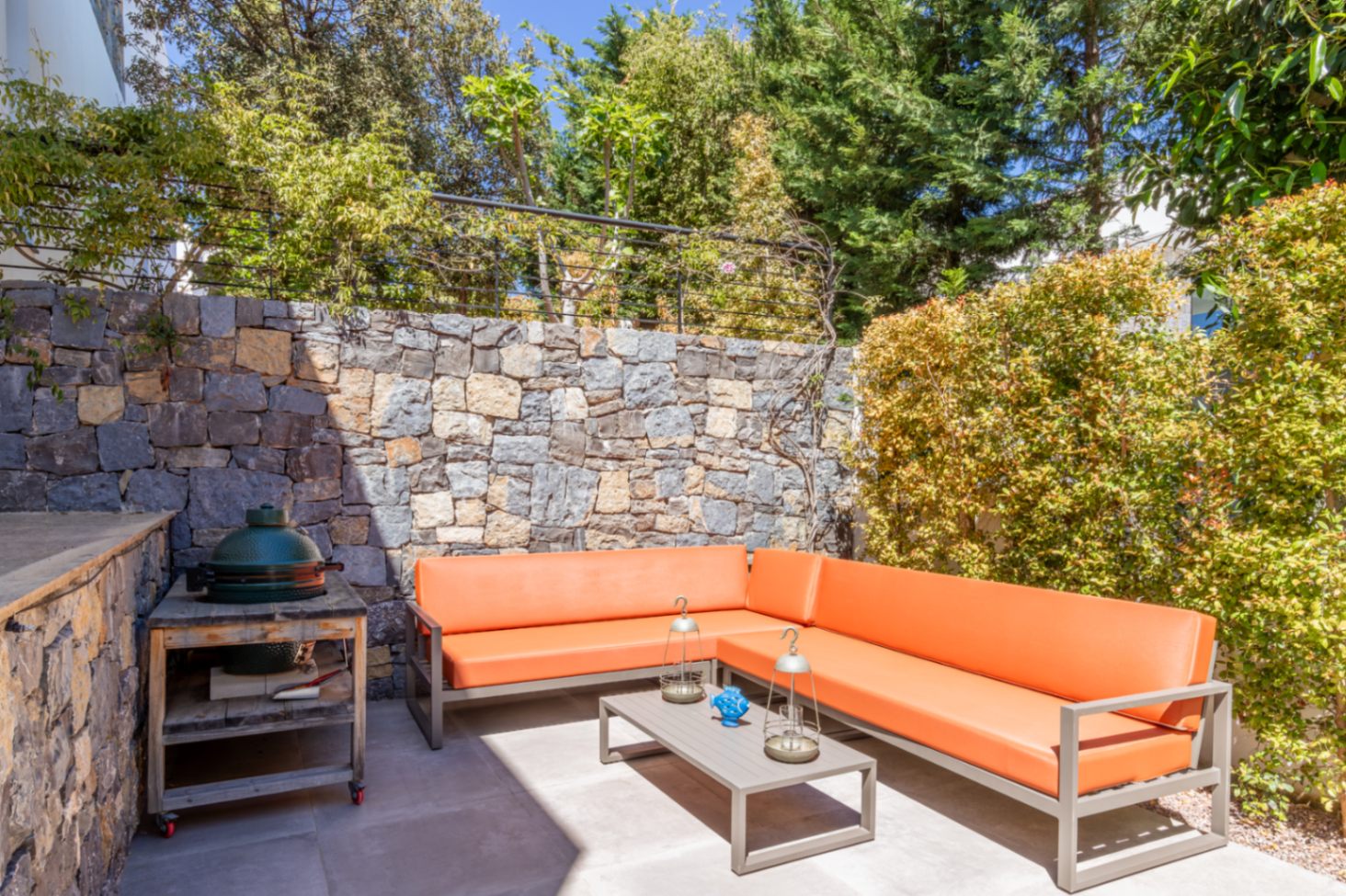 Buitenlounge met oranje hoekbank, keramische kamado-barbecue en natuurstenen muur, omgeven door volwassen mediterrane beplanting.