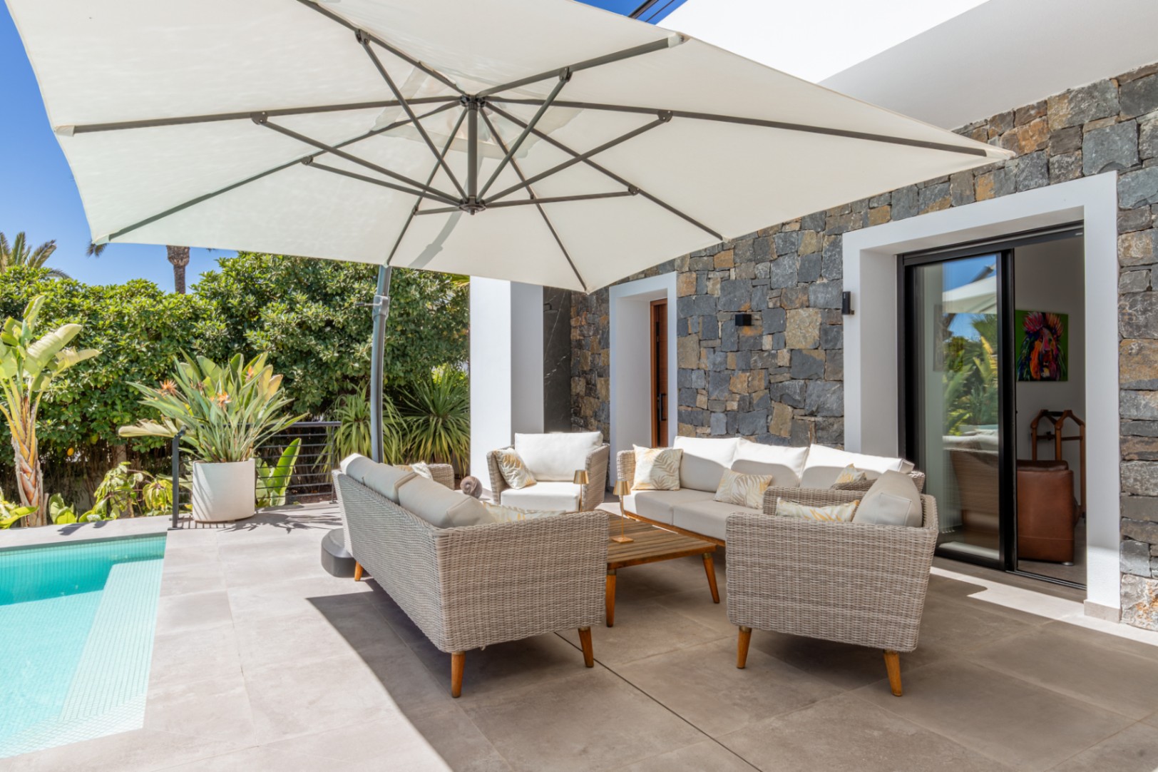 Buitenlounge met rieten meubilair en een grote parasol op een betegeld terras naast het zwembad, afgewerkt met een natuurstenen gevel.
