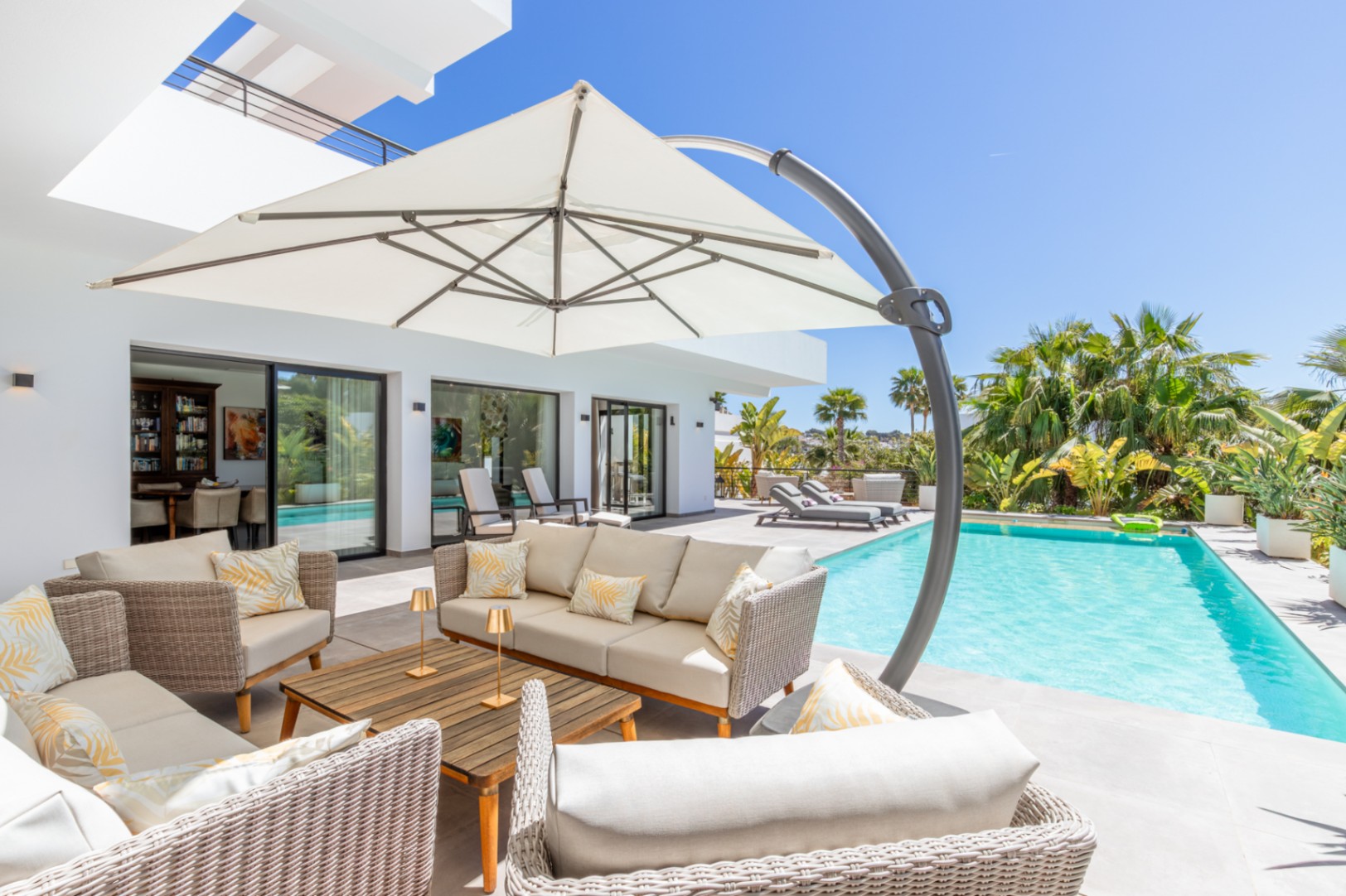 Buitenlounge met wicker zitmeubilair en grote parasol bij het zwembad, met directe toegang tot de woning via grote glazen schuifdeuren.