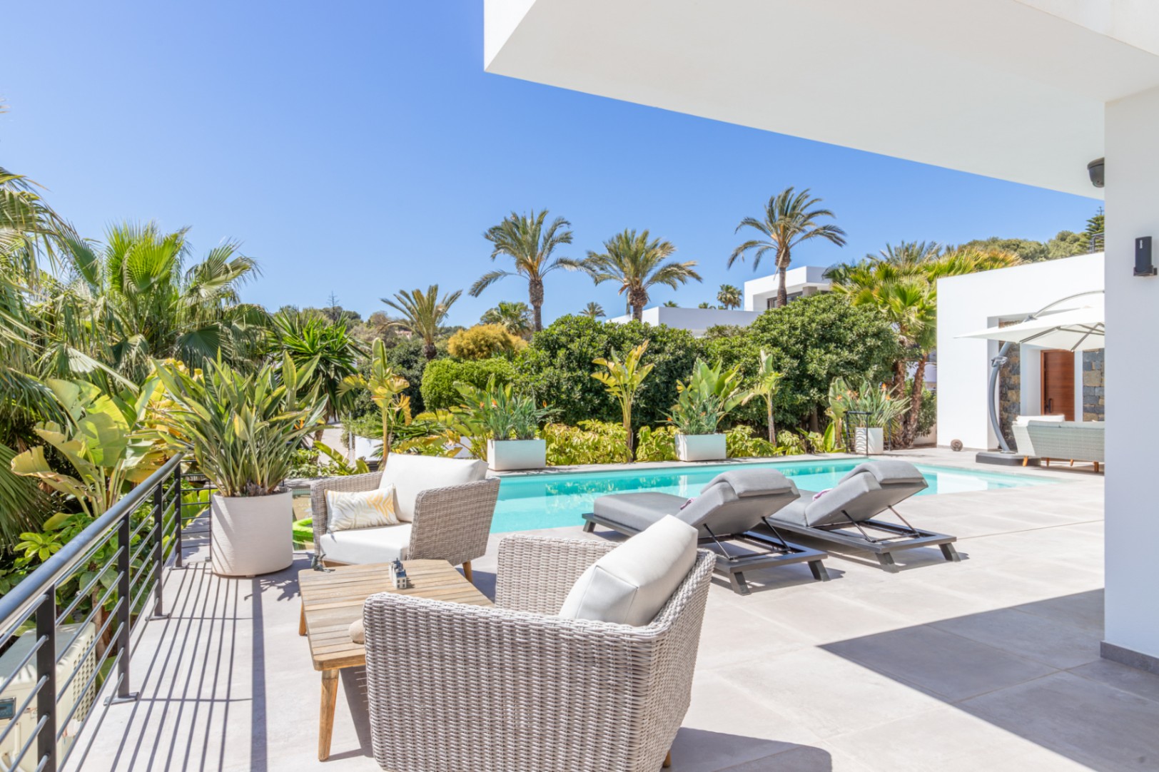 Buitenterras met rotan meubilair en ligbedden bij het zwembad, omgeven door een volgroeide mediterrane tuin met palmbomen en veel privacy.