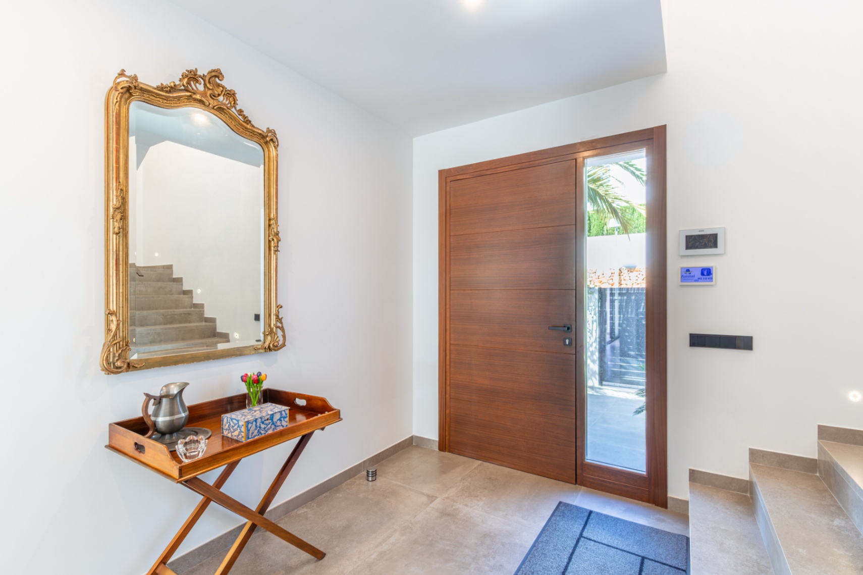 De hal is voorzien van een grote houten voordeur met zijlicht, een rijk gedecoreerde gouden spiegel en een moderne stenen vloerafwerking.
