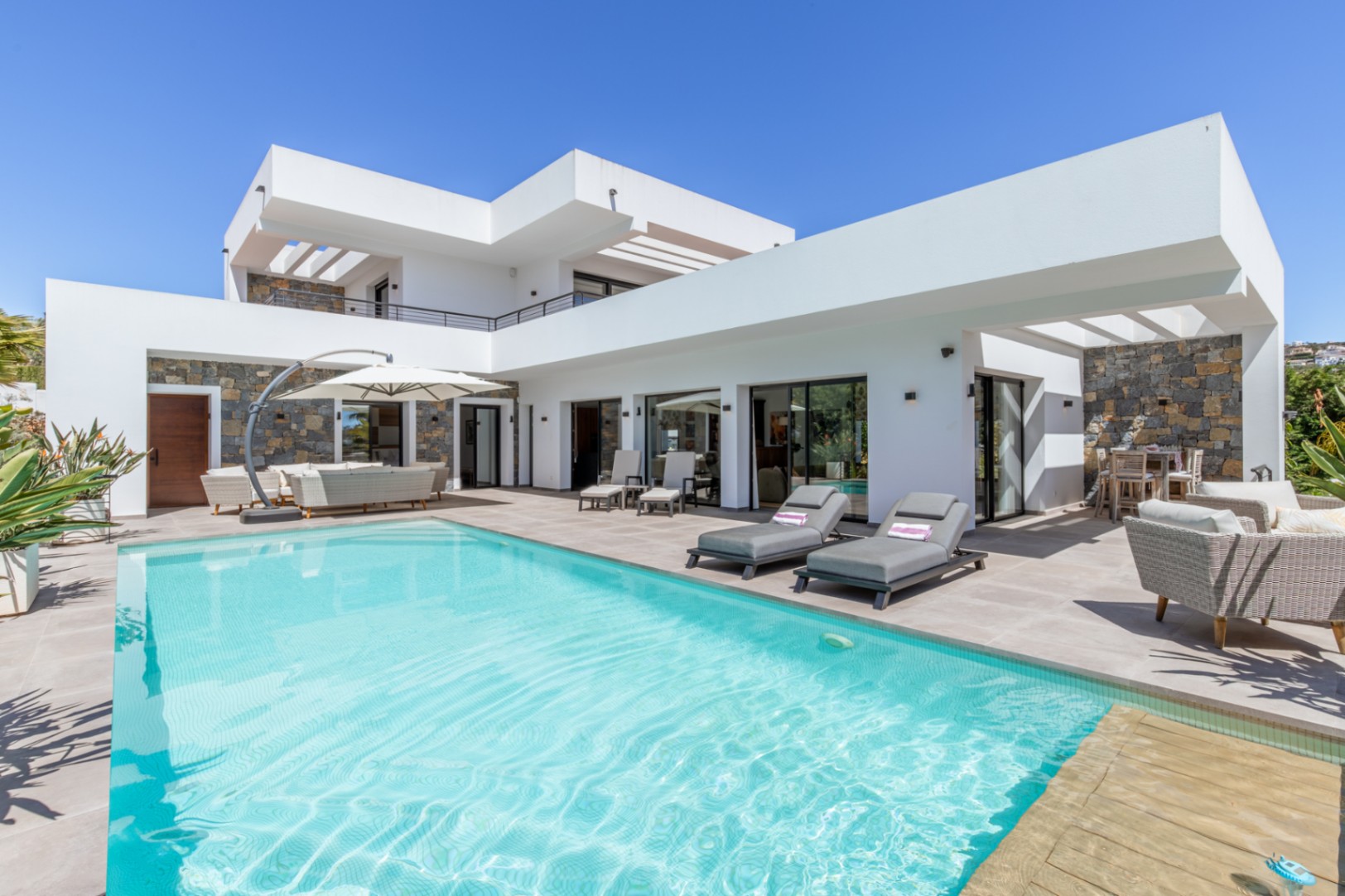 Exterieur van de villa met een groot zwembad, ruim terras en moderne witte architectuur afgewerkt met natuurstenen muren en strakke lijnen.