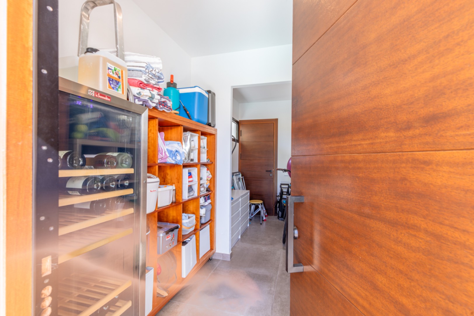 Functionele bijkeuken en voorraadkast met houten rekken, een geïntegreerde wijnkoeler en directe toegang via hoogwaardige houten deuren.
