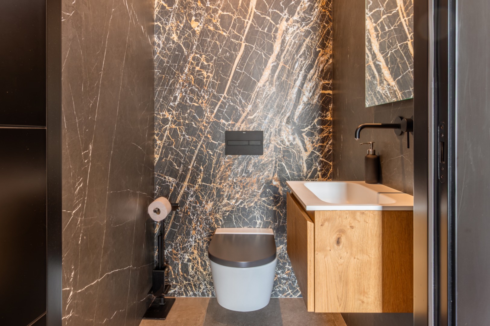 Gastentoilet met een wand van donker marmer met gouden aders, hangend toilet en een eikenhouten wastafelmeubel met matzwarte kranen.