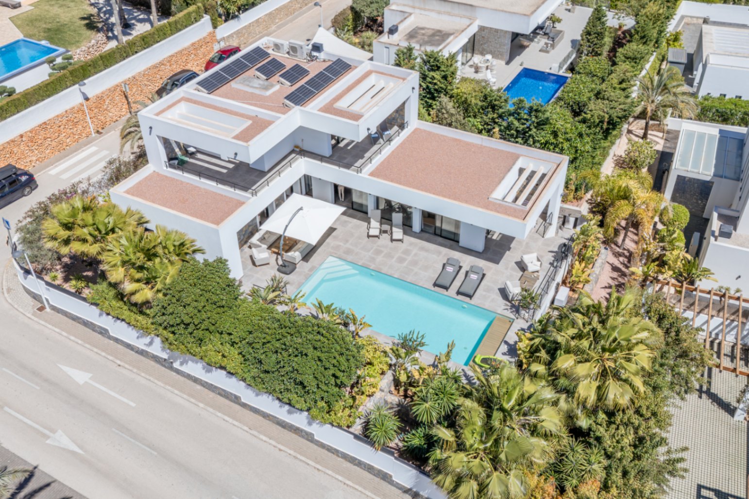 Luchtfoto van de moderne villa met zonnepanelen, een rechthoekig zwembad en ruime terrassen omgeven door volgroeide mediterrane beplanting.