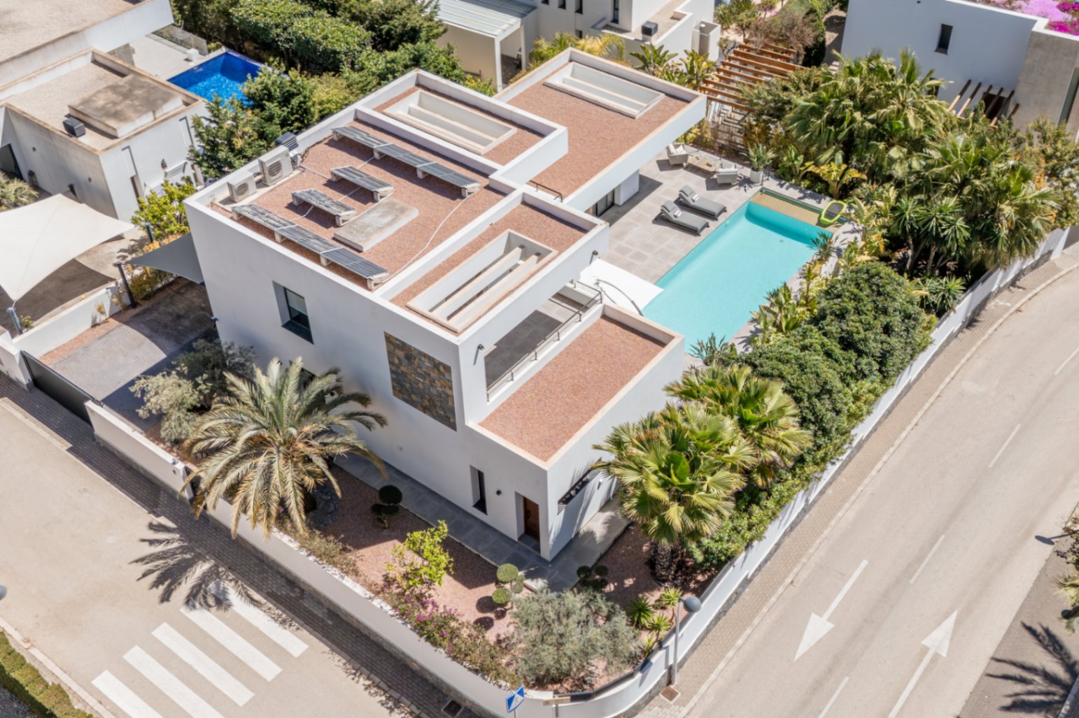 Luchtfoto van de moderne villa met zonnepanelen op het dak, een privézwembad met terras en volgroeide mediterrane beplanting op een hoekperceel.
