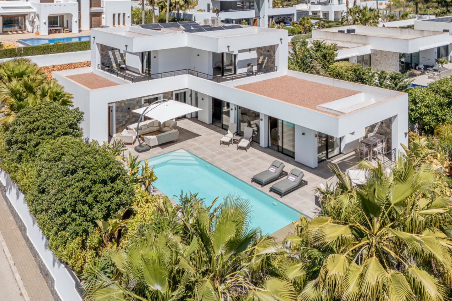Luchtfoto van de moderne villa met zwembad, zonnepanelen op het dak en ruime terrassen omgeven door volgroeide mediterrane beplanting.