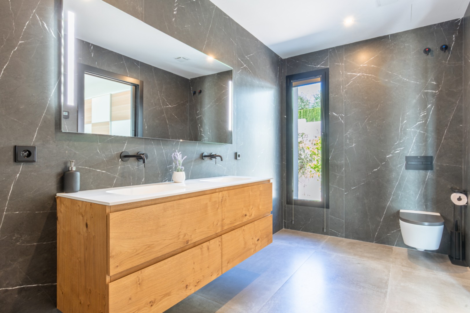 Moderne badkamer met donkere marmerlook wandtegels, houten dubbel wastafelmeubel en matzwarte kranen. Voorzien van een hangend toilet en een grote spiegel met geïntegreerde LED-verlichting.