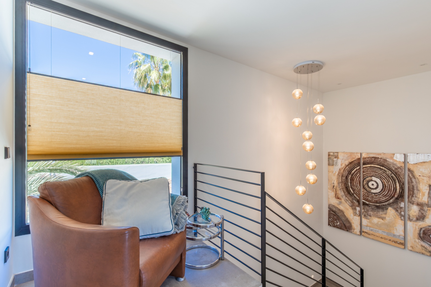 Overloop op de bovenverdieping met lederen fauteuil, moderne hanglamp met bollen en een groot raam met uitzicht op de tuin en een palmboom.