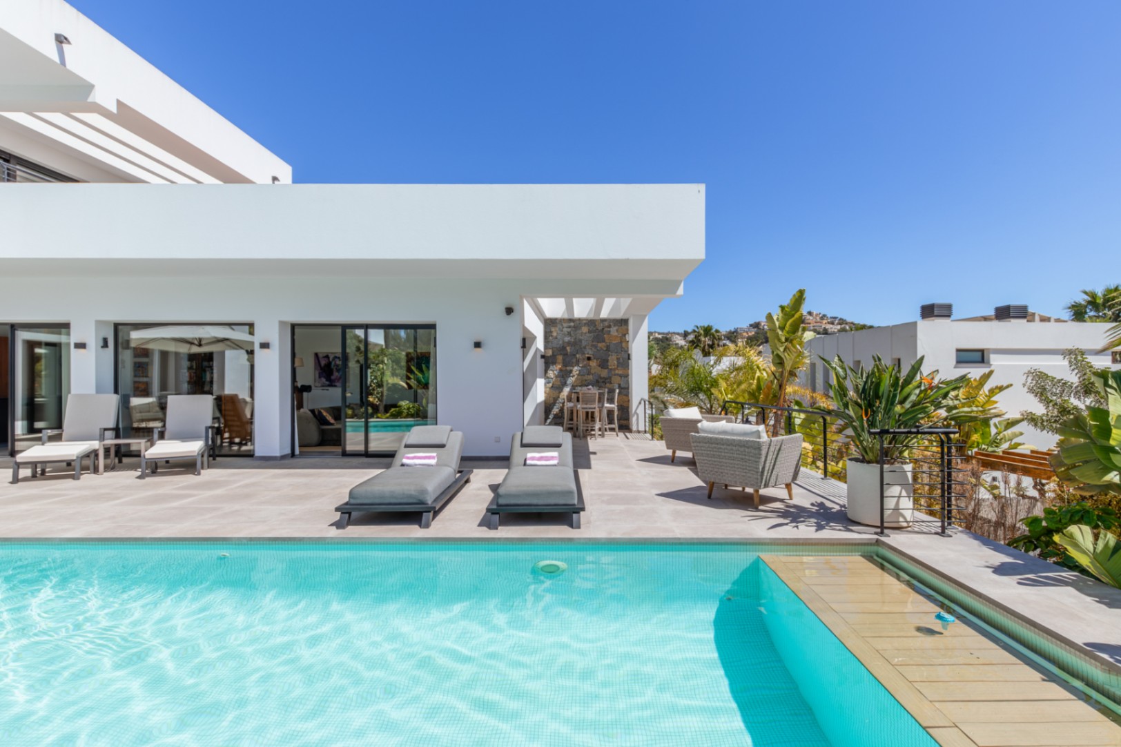 Ruim terras met turquoise betegeld zwembad, ligbedden en een eethoek onder de pergola, omgeven door moderne witte architectuur en mediterrane beplanting.