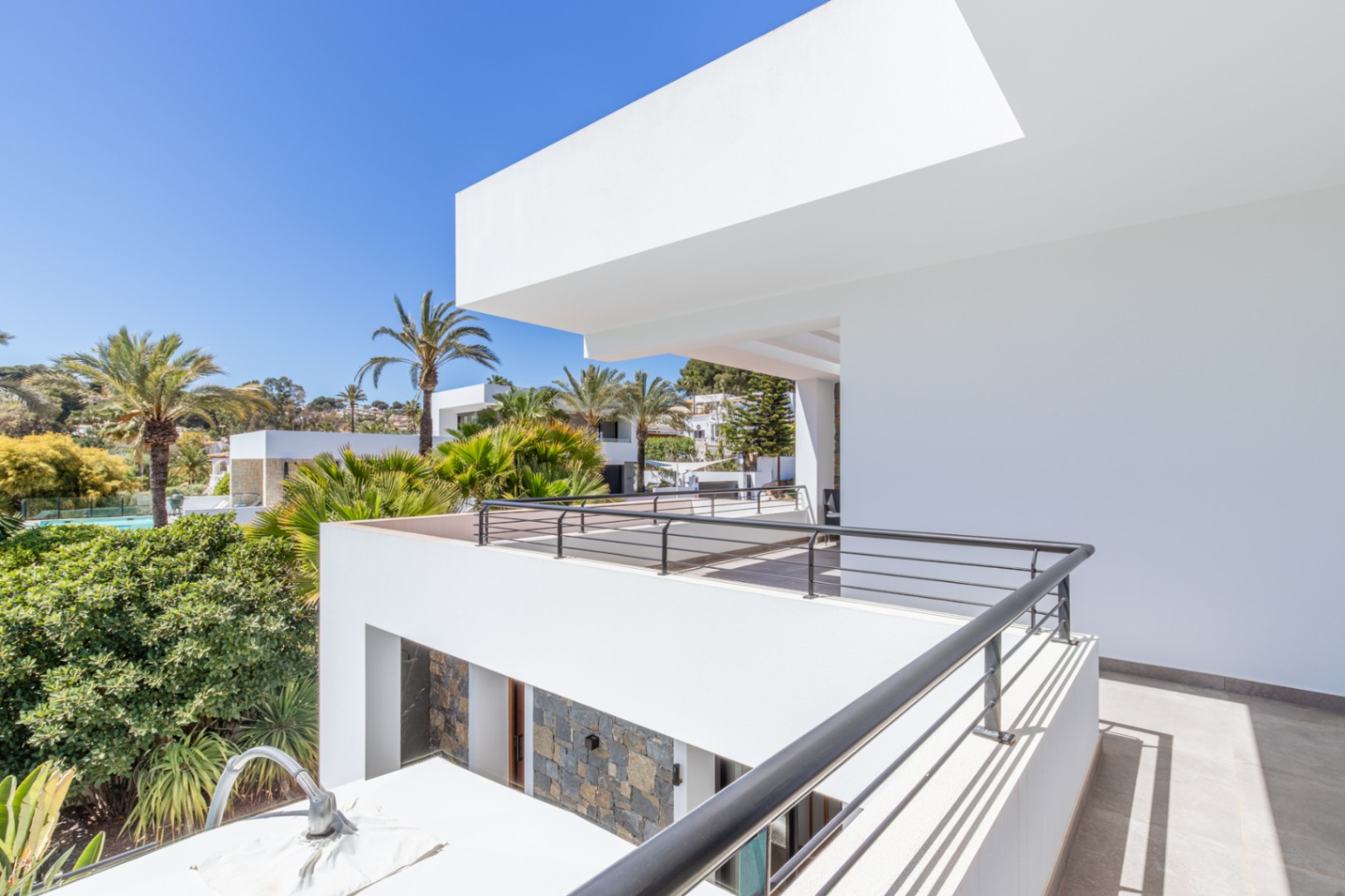 Terras op de bovenverdieping met zwart metalen balustrade en uitzicht op de mediterrane tuin. De moderne witte gevel is voorzien van natuurstenen details.