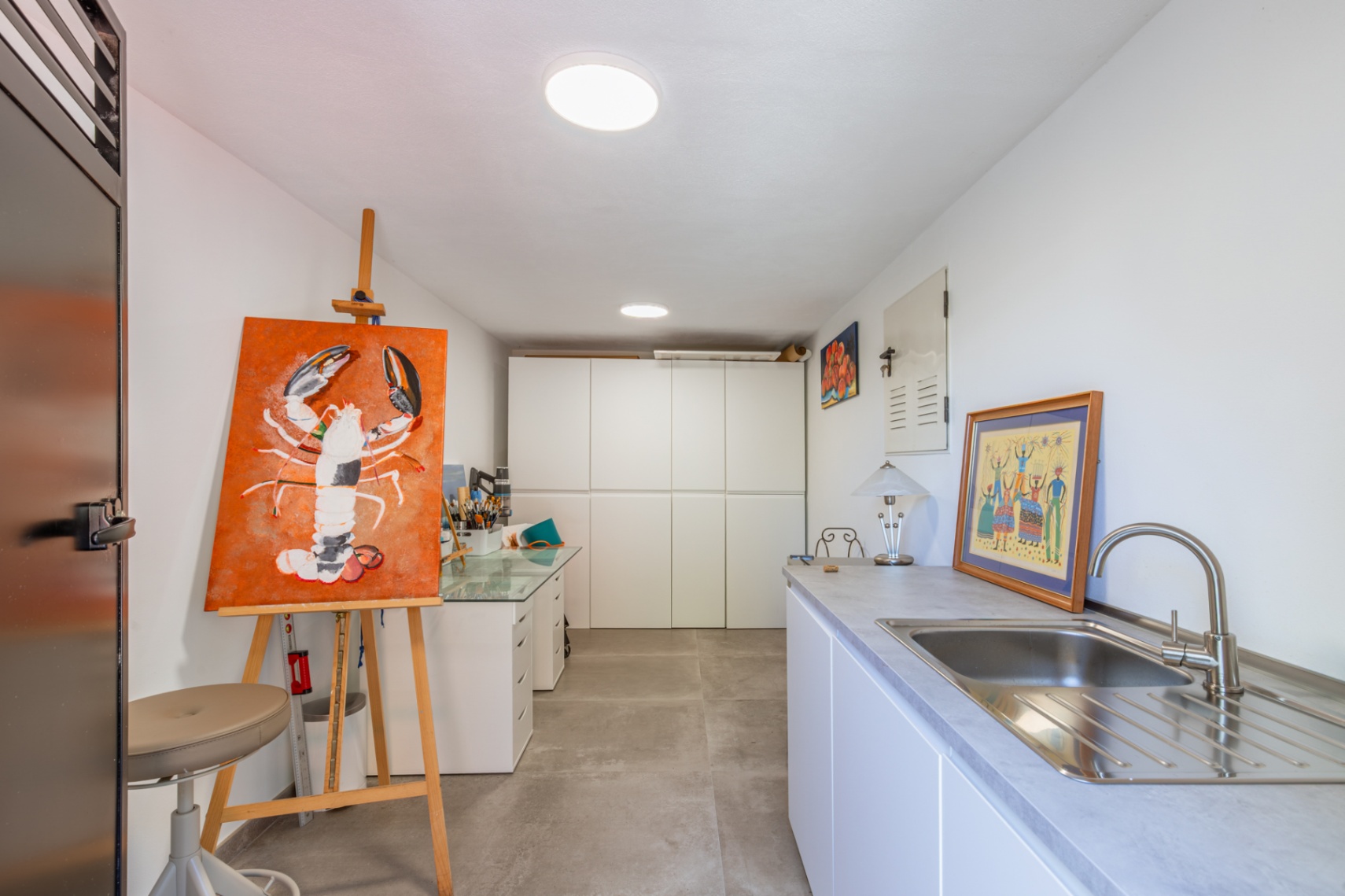 Veelzijdige studio- of bijkeuken met witte inbouwkasten, een roestvrijstalen spoelbak en een artistieke werkplek op grijze porseleinen tegels.