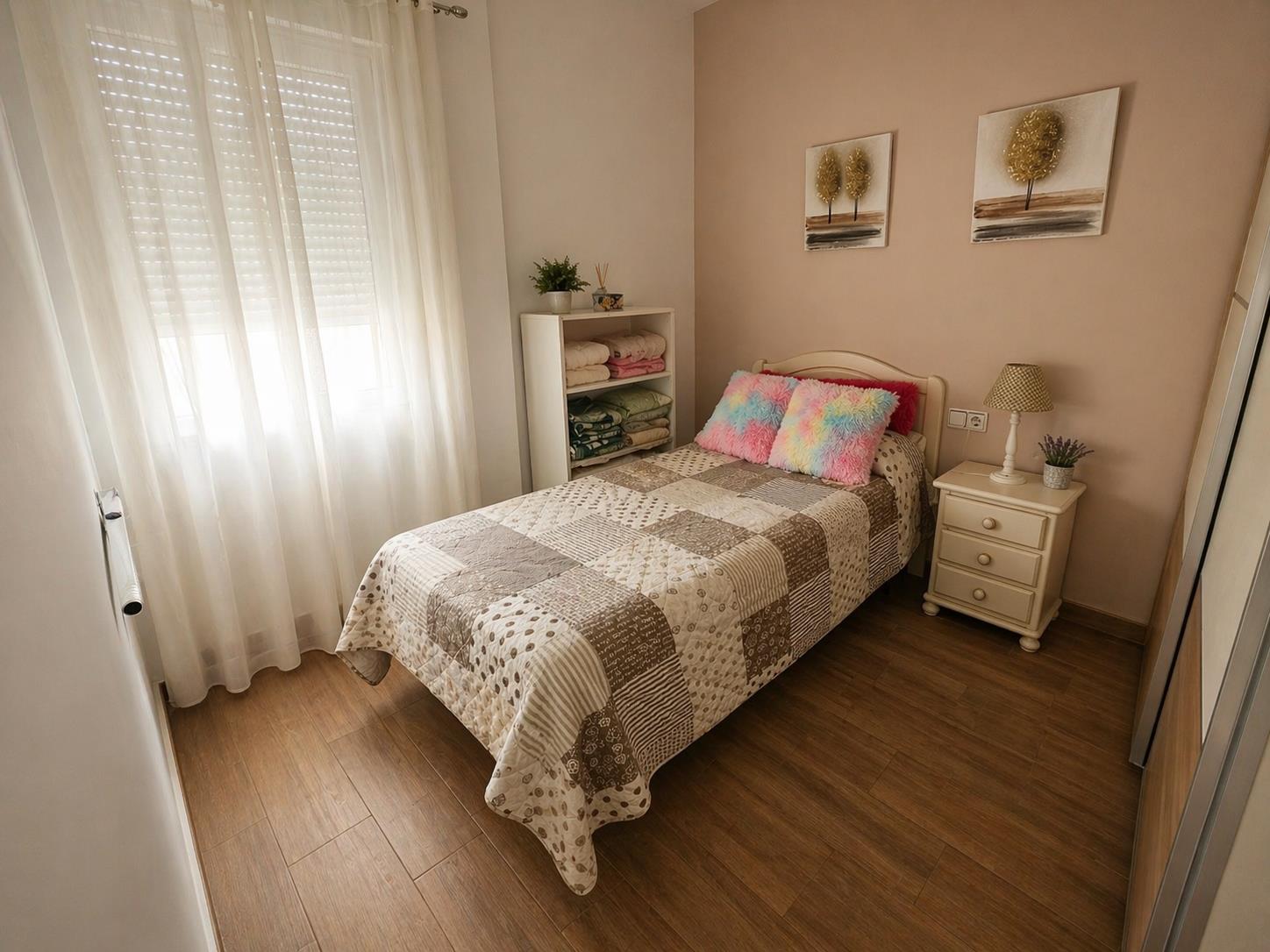 Eenpersoonsslaapkamer met houtlook vloertegels, raam met rolluiken en witte gordijnen, en een accentmuur in aardetinten.
