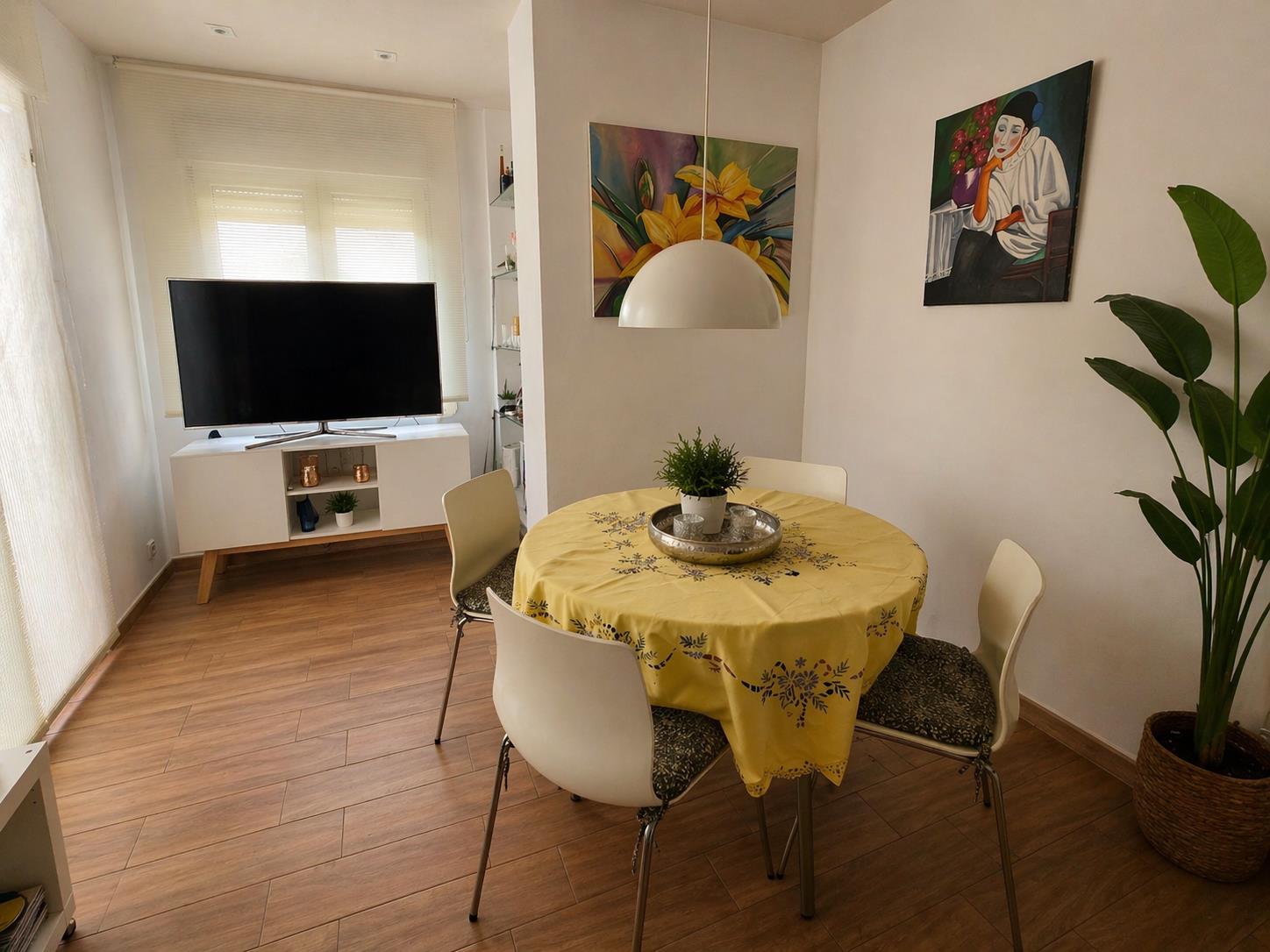 Eethoek met een ronde tafel, witte stoelen en een hanglamp, gelegen in een lichte woonkamer met een vloer in houtlook en een tv-meubel.