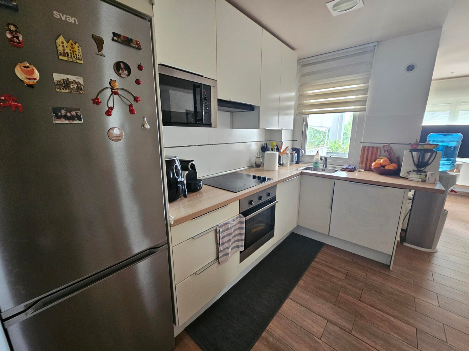 Functionele L-vormige keuken met houtlook werkbladen, inbouwapparatuur en een buitenraam. De keramische tegelvloer imiteert de warmte van hout.