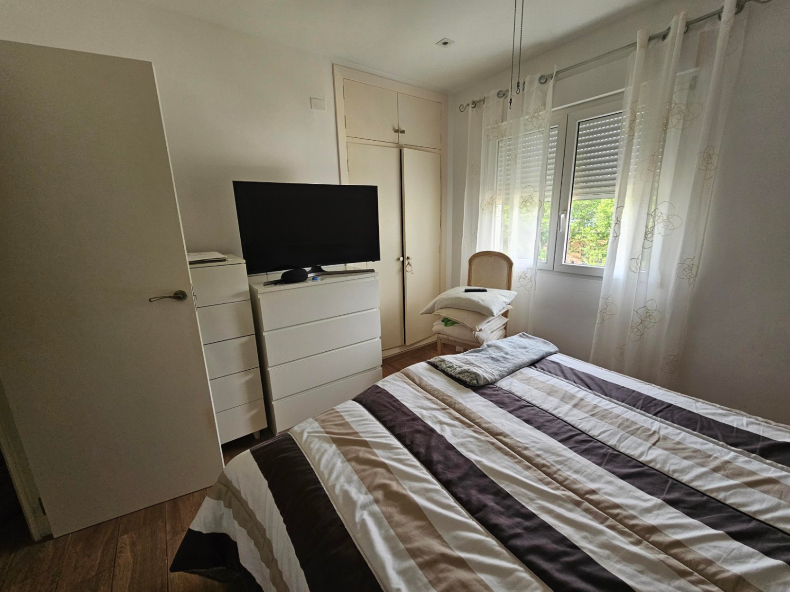 Functionele slaapkamer met inbouwkasten, witte ladekasten en een raam met rolluiken dat natuurlijk licht binnenlaat op de laminaatvloer.